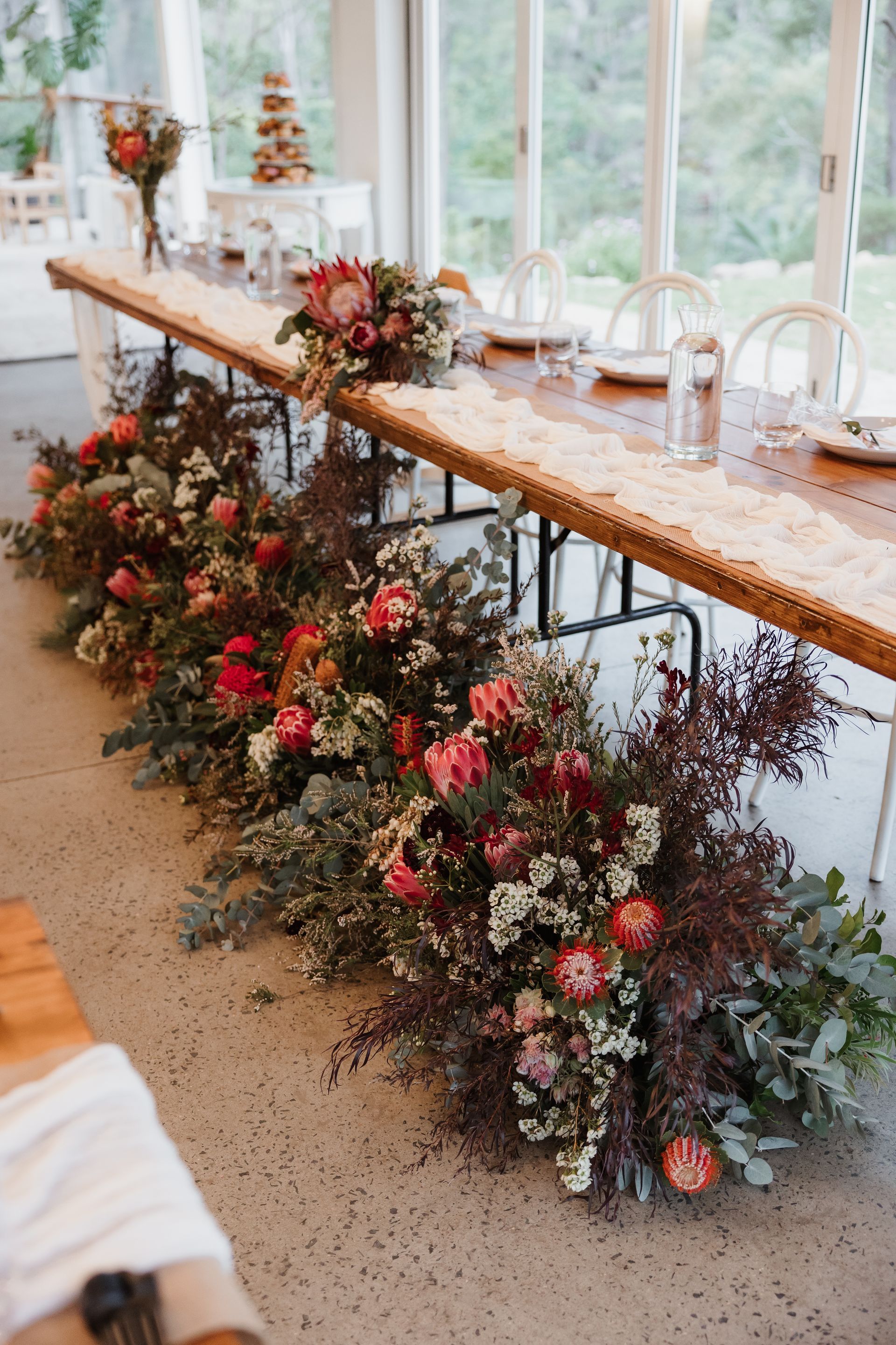 Long wooden table decorated with floral arrangement. Protea, red flowers, and greenery.