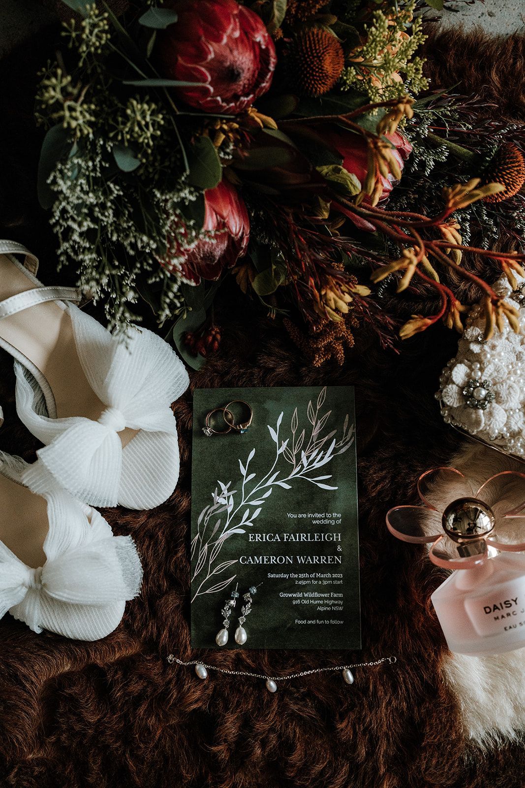 Wedding invitation with floral bouquet, shoes, jewelry, and perfume on a textured surface.