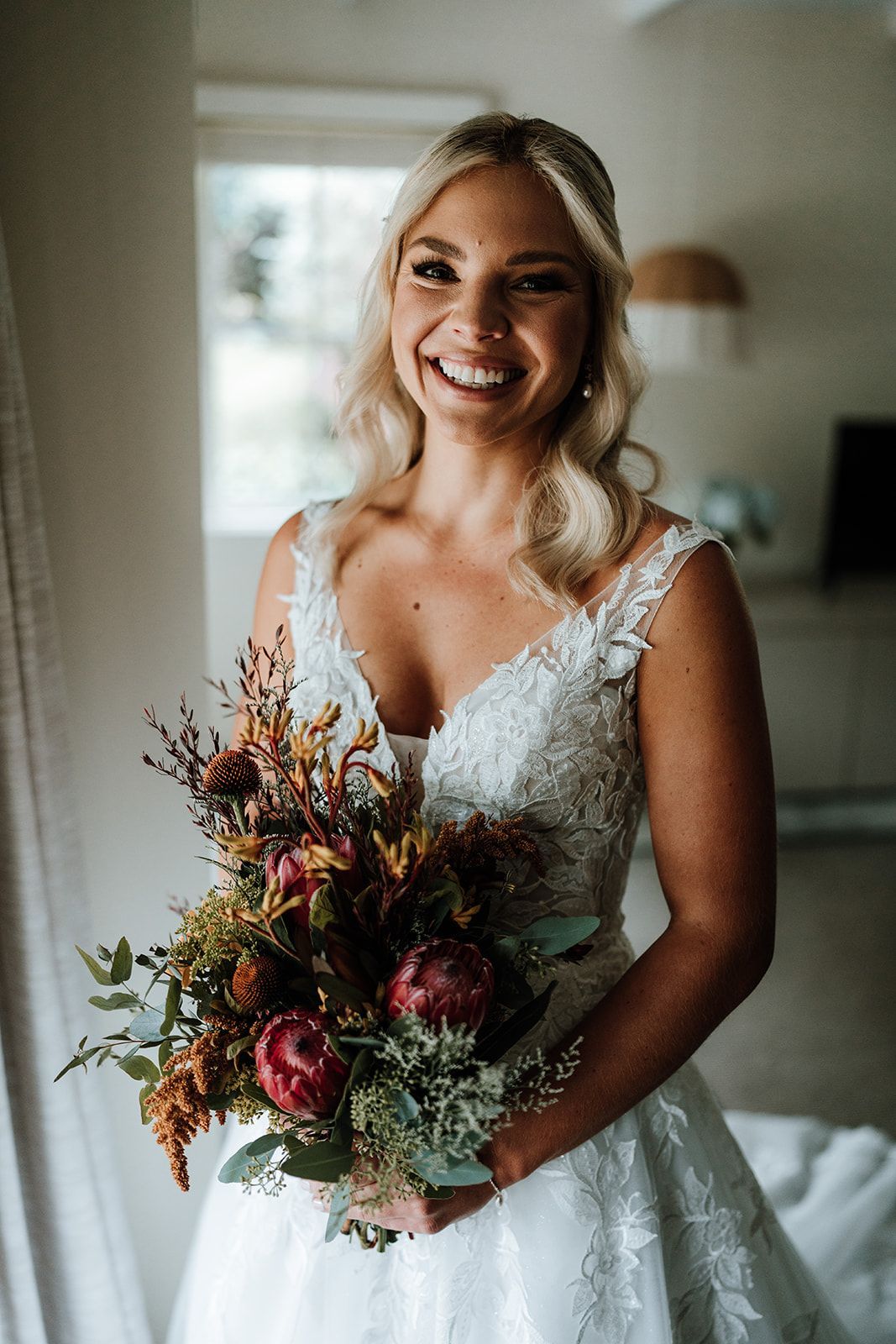 Bride in a white lace wedding dress smiles holding a bouquet of flowers indoors.