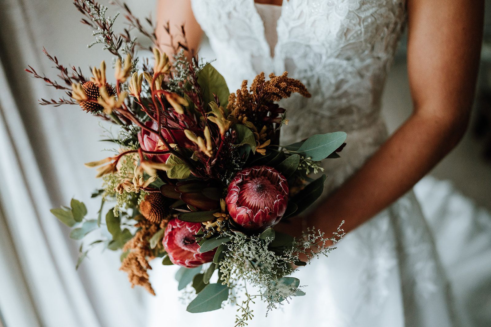 Bride holding rustic bouquet with protea flowers, against a white lace dress.