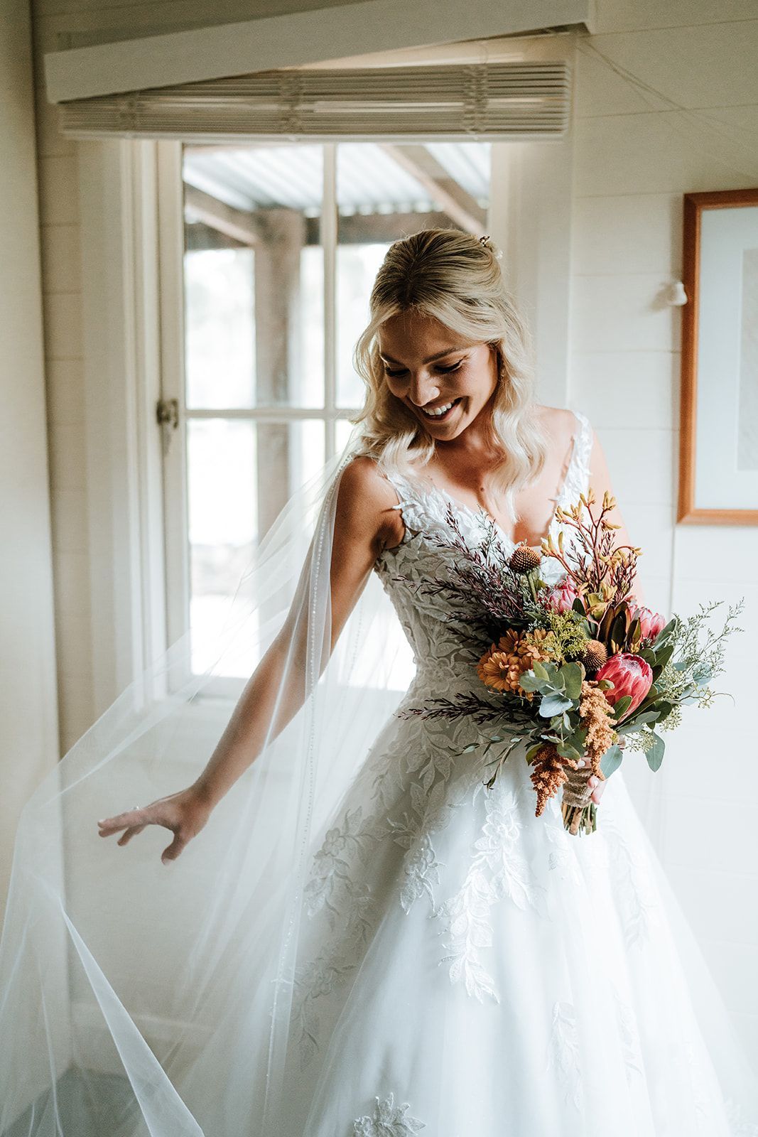 Bride in white wedding dress, holding bouquet, smiling, near window.