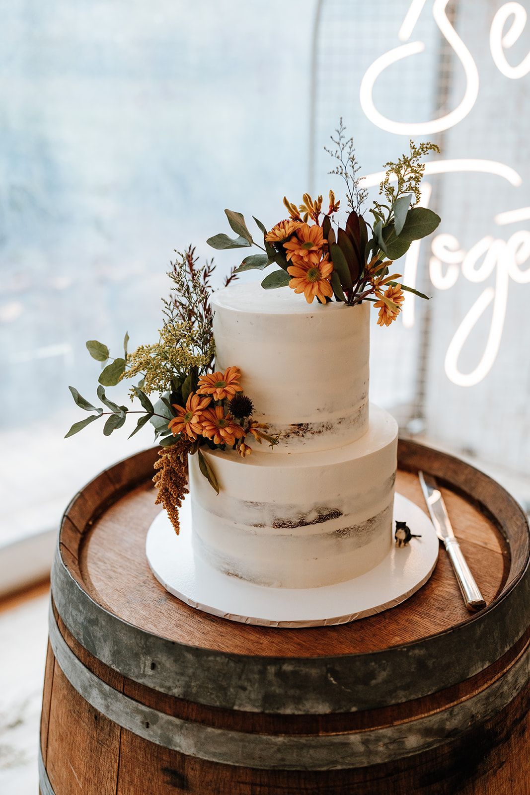 Two-tiered wedding cake with floral arrangement on a wooden barrel, next to a knife.