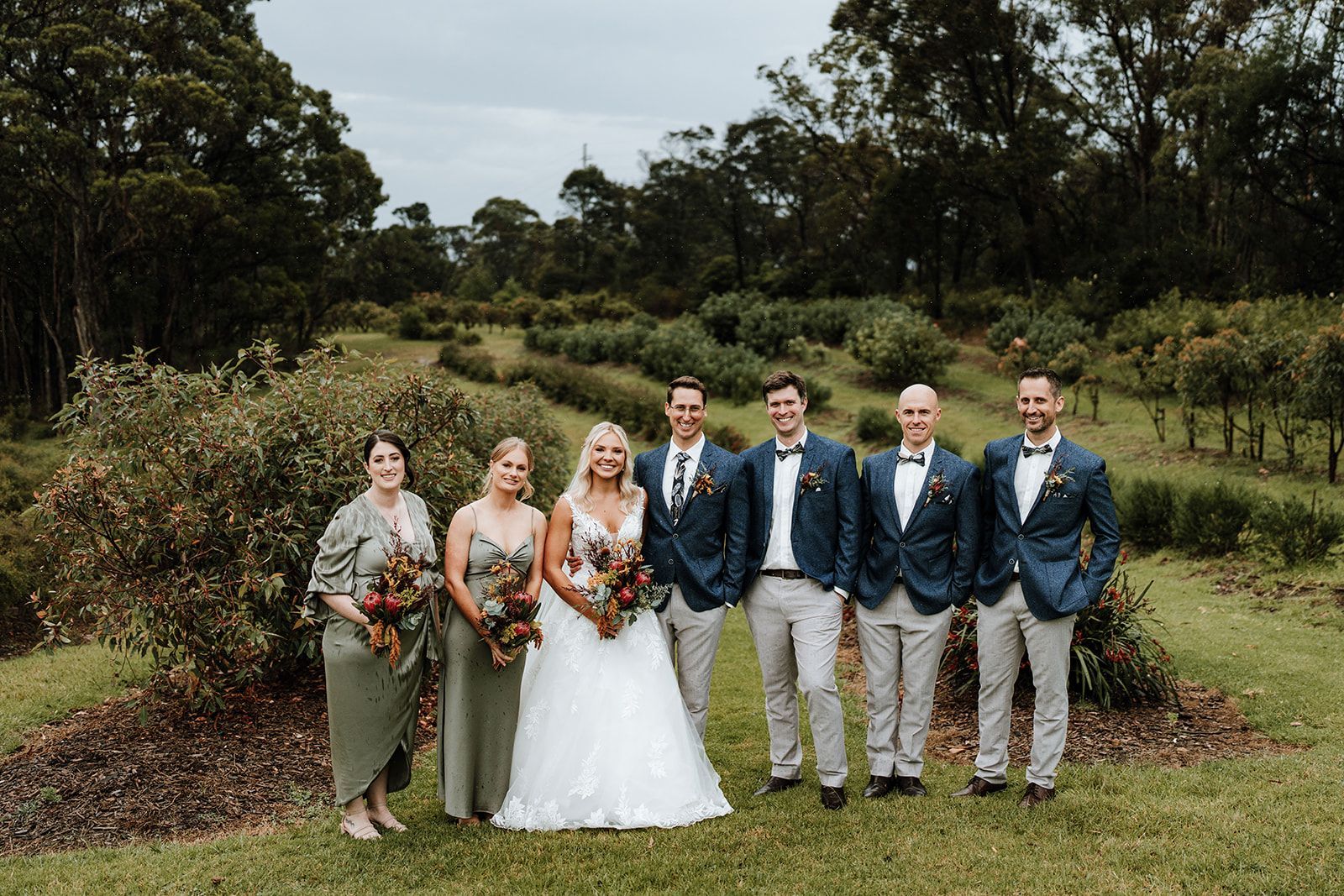 Wedding party poses outdoors on a grassy lawn; bride, bridesmaids, and groomsmen in formal attire with bouquets.