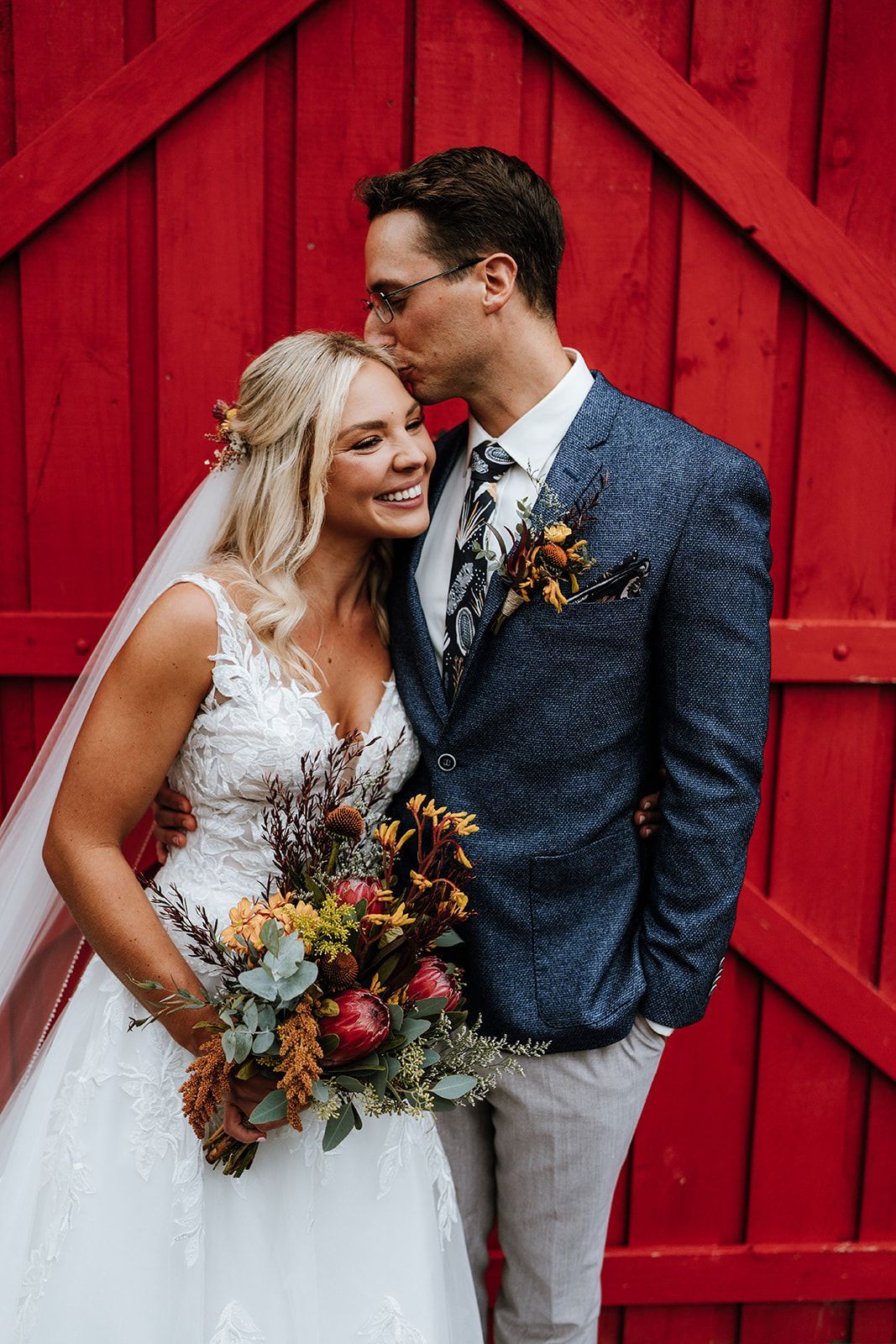 Bride and groom pose in front of a red door, smiling. Groom kisses bride's forehead, holding bouquet.