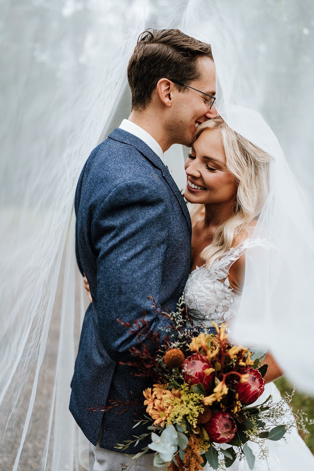 Bride and groom embrace, veiled by fabric, near foliage. Groom kisses bride’s head; bride holds flowers.