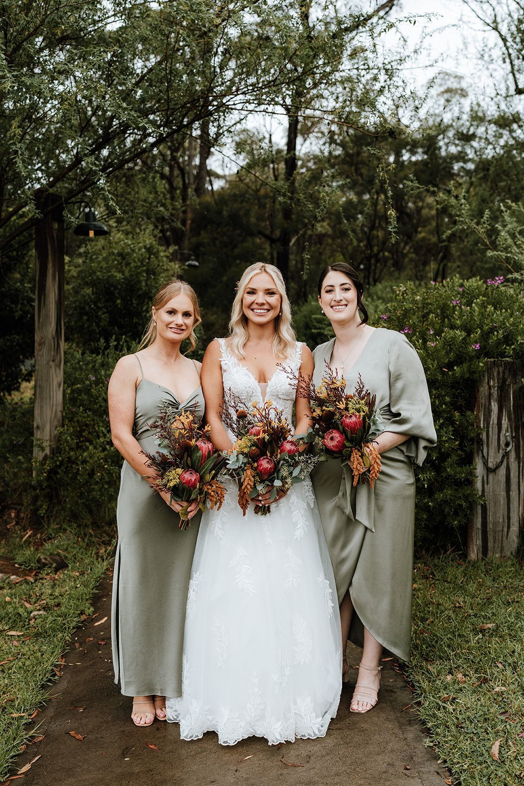Bride in white dress with two bridesmaids in olive green dresses, holding bouquets outdoors.