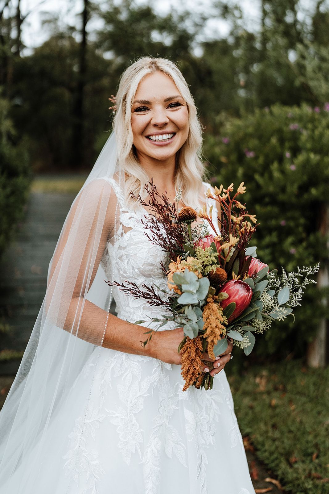 Bride in white dress, holding bouquet, smiling at camera. Outdoors with greenery in background.