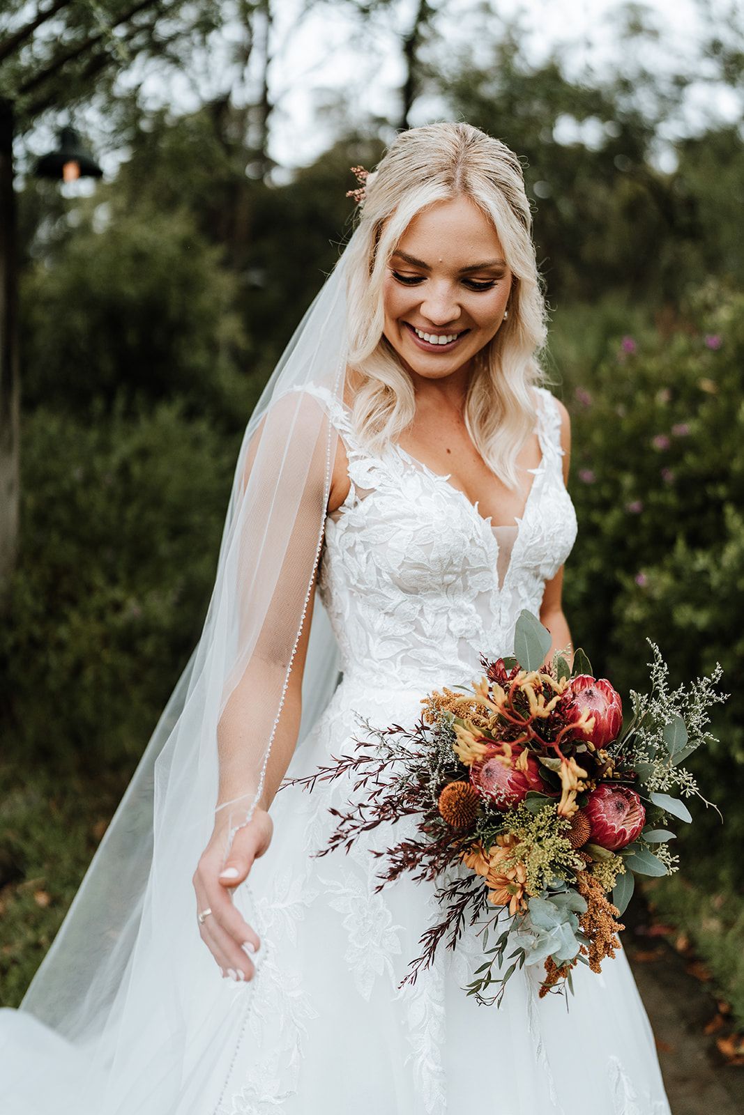 Blonde bride smiles, holding bouquet, wearing a white lace wedding dress with a veil, outdoor setting.