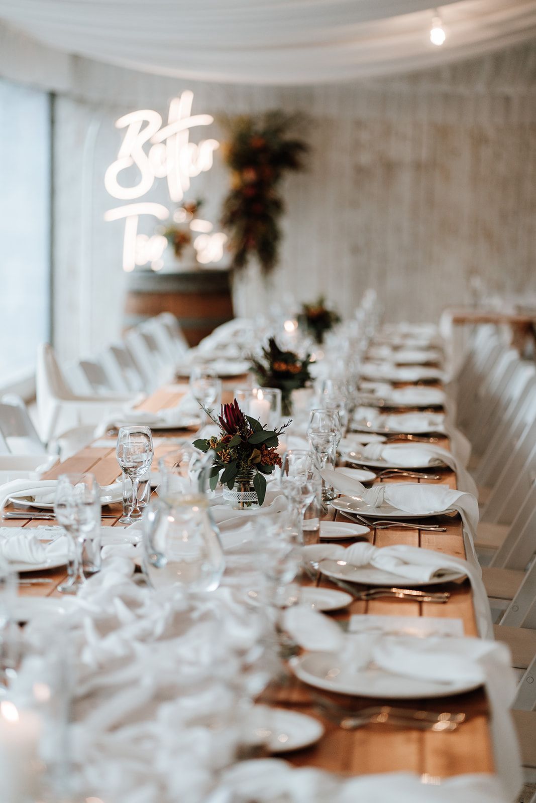 Long wooden table set for an event, white decor, floral centerpieces, neon sign: 