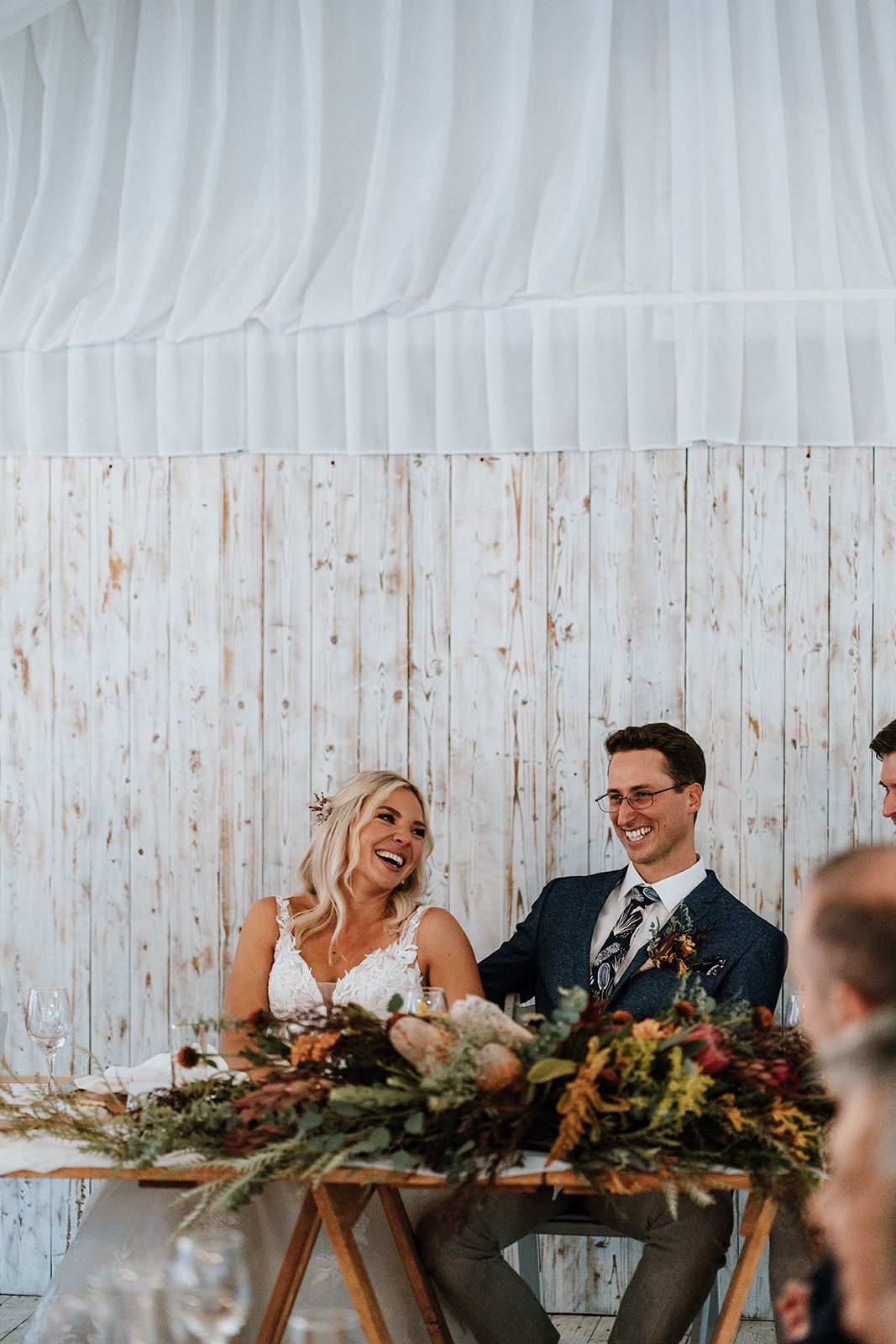 Wedding reception: Bride and groom laughing at a table with floral arrangement, white wood backdrop.