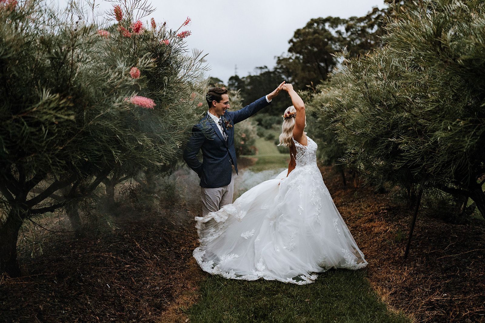 A couple dances in a garden path; the bride wears a white dress, the groom a blue blazer.