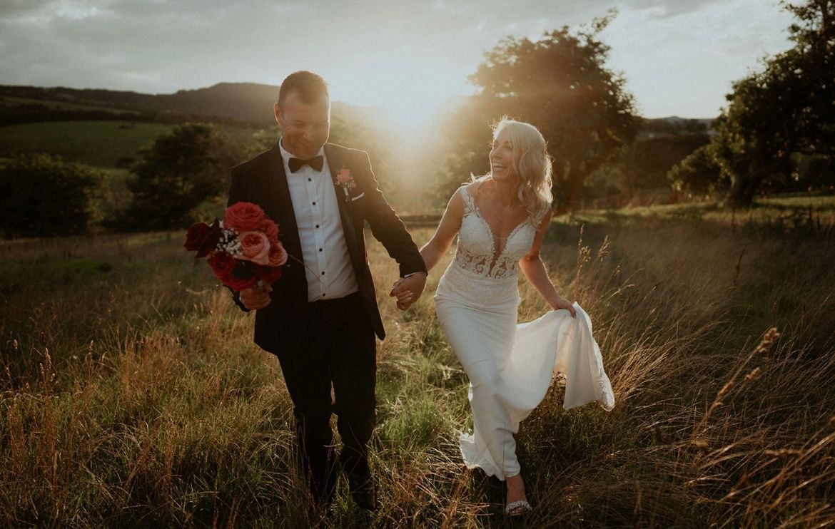 Bride and groom hold hands walking through a field at sunset, wearing a dress and a tuxedo.