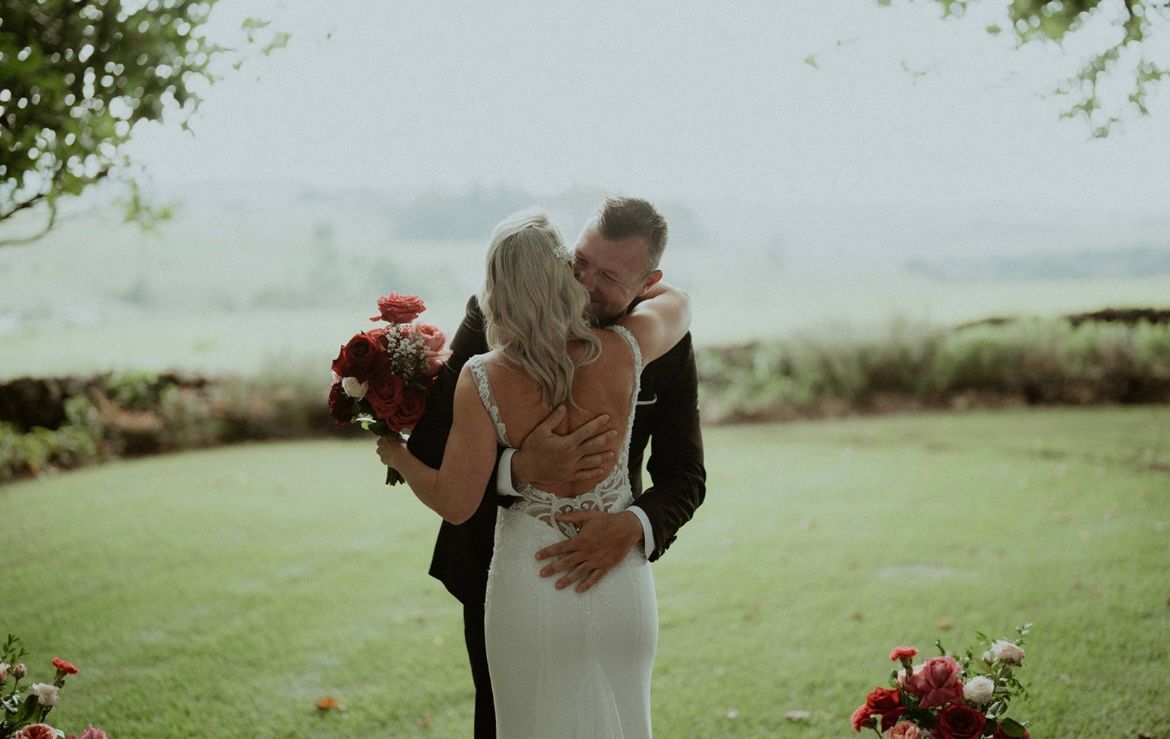 Newlyweds embrace; bride in white dress holds bouquet, groom in a black suit, outdoor wedding.