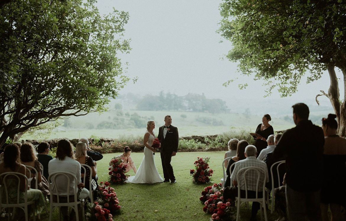 Wedding ceremony with couple standing at altar, red floral arrangements, and seated guests on a grassy lawn.