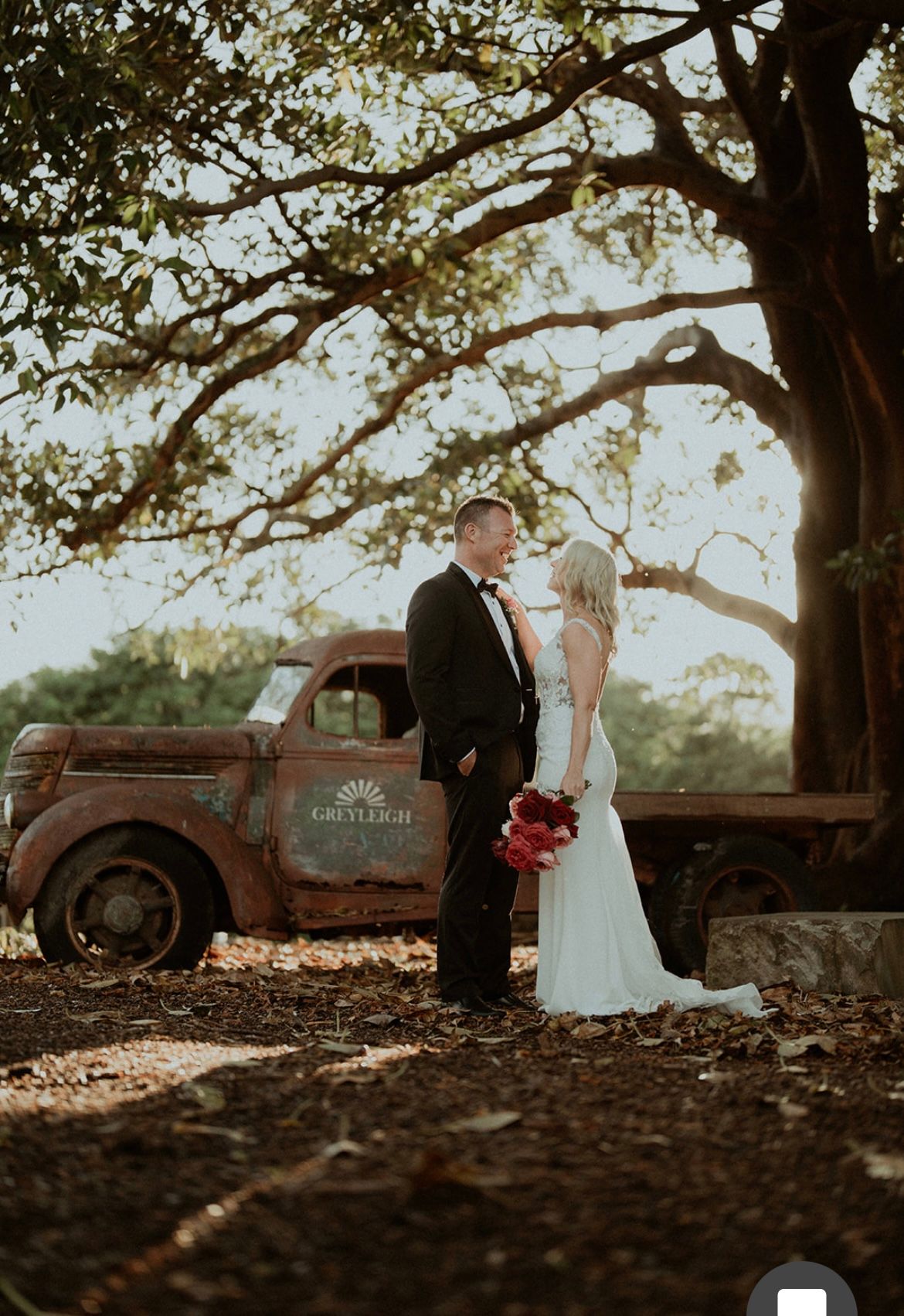 Wedding couple by a vintage truck under a large tree. The bride wears a white dress and the groom a suit.