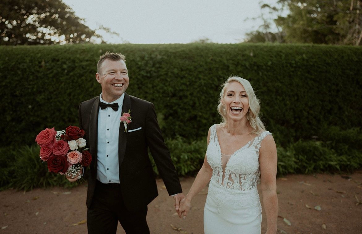 Newlyweds holding hands, laughing, walking, groom in tuxedo, bride in white dress, against green hedge.