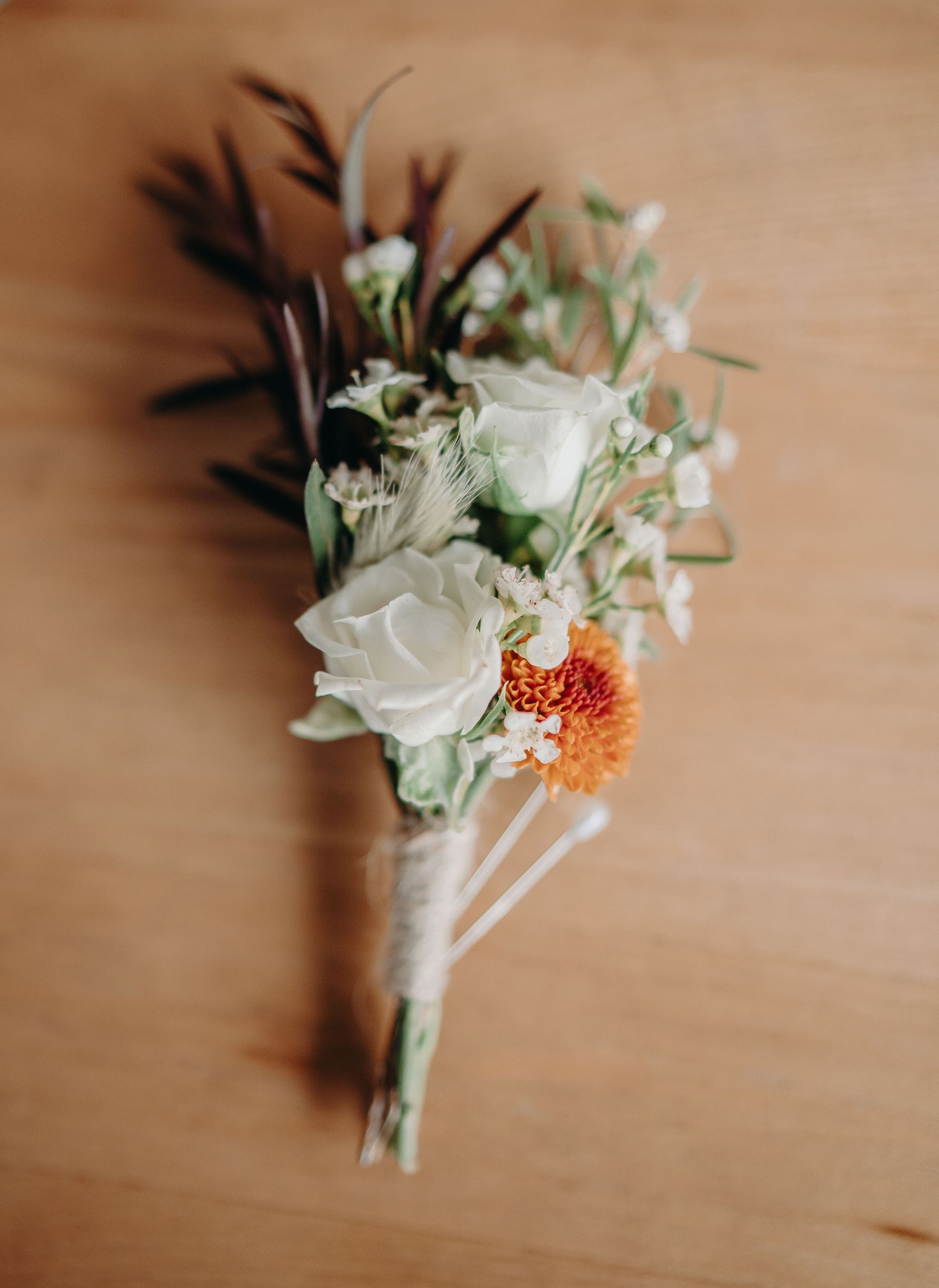 Boutonniere with white roses, orange flower, and greenery, tied with twine, on a wood surface.
