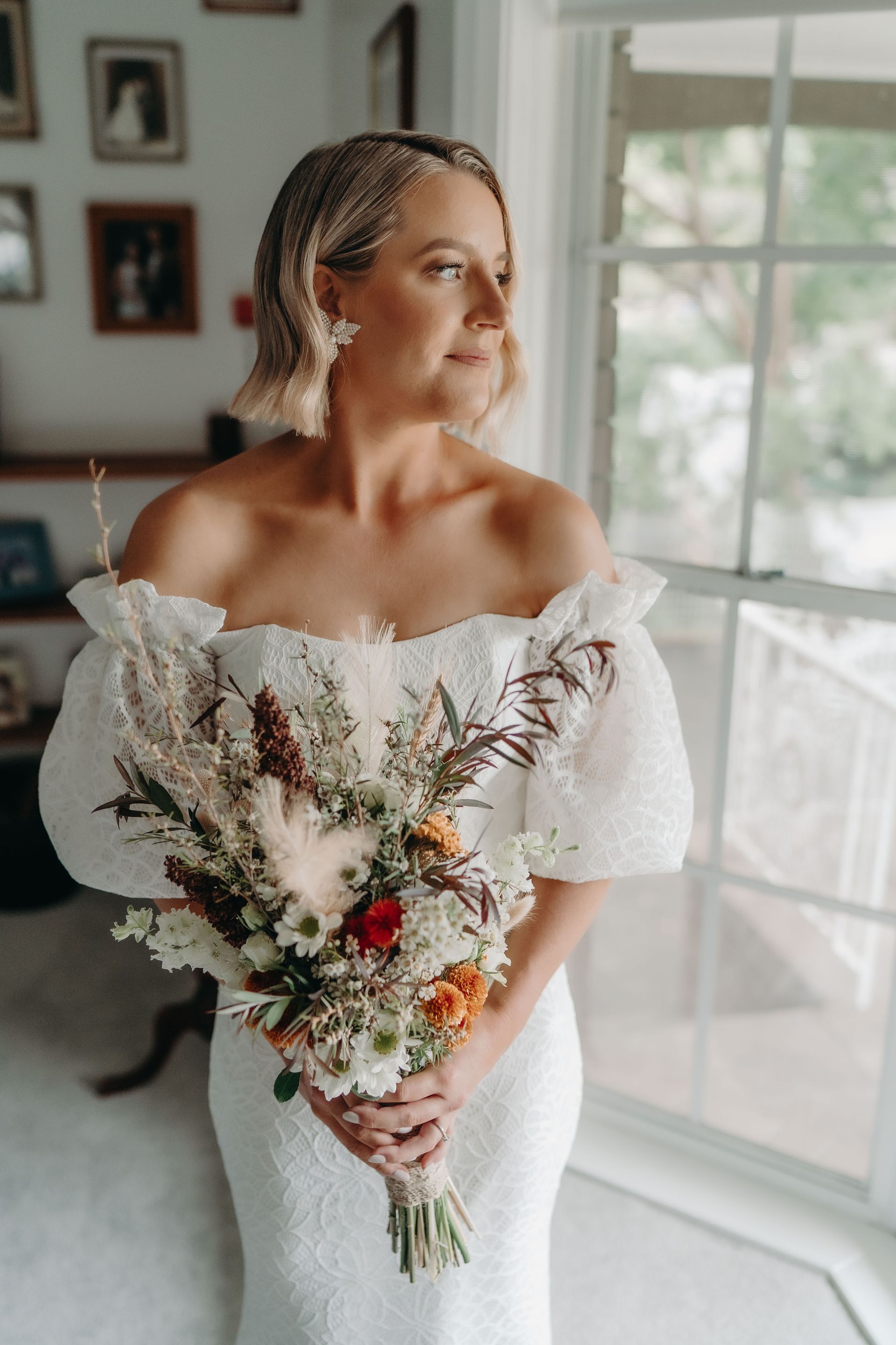 Bride in off-shoulder dress, holding a bouquet, looks out a window.