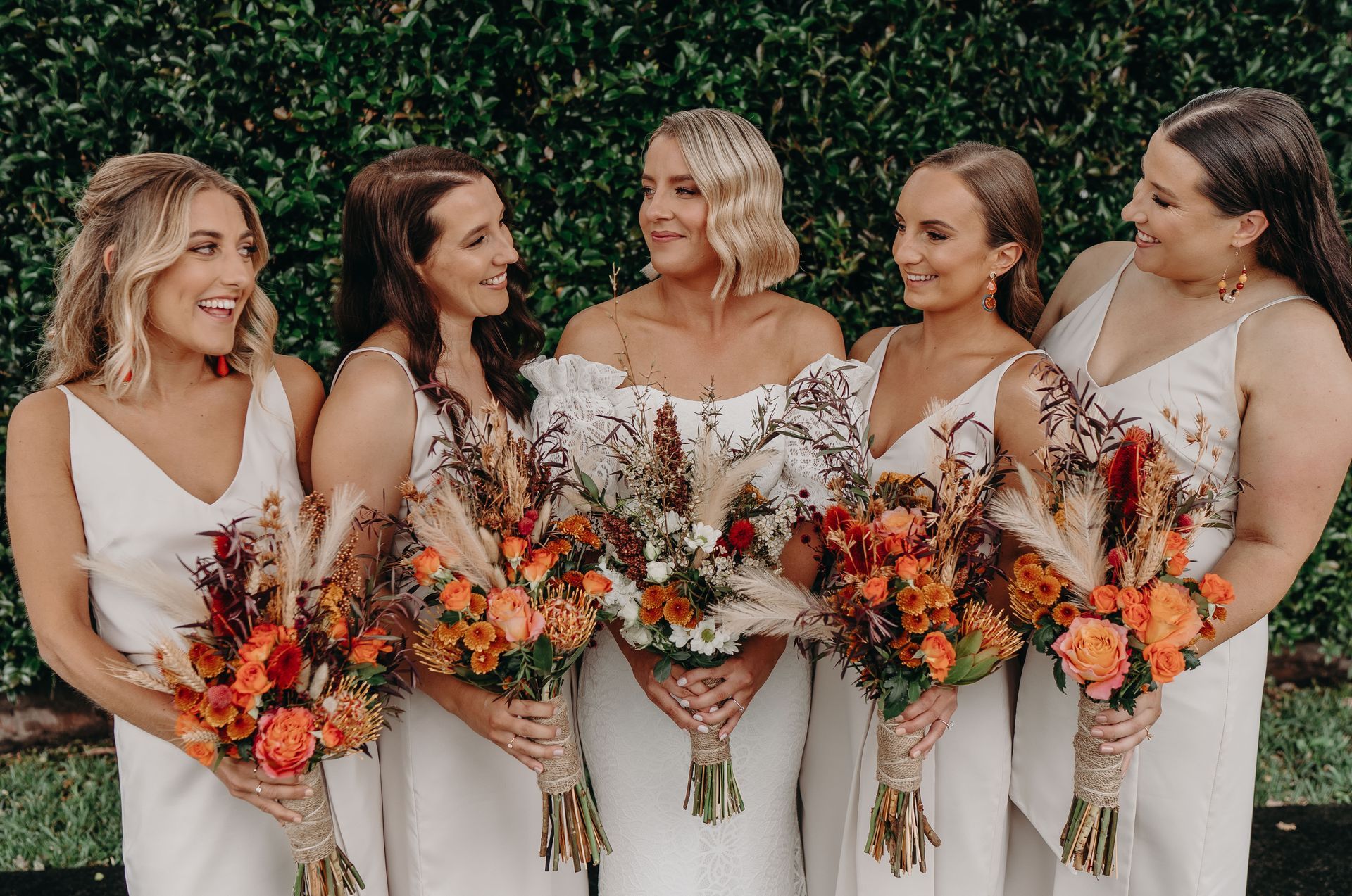 Bridesmaids in cream dresses hold bouquets with orange flowers, smile, and stand outdoors.