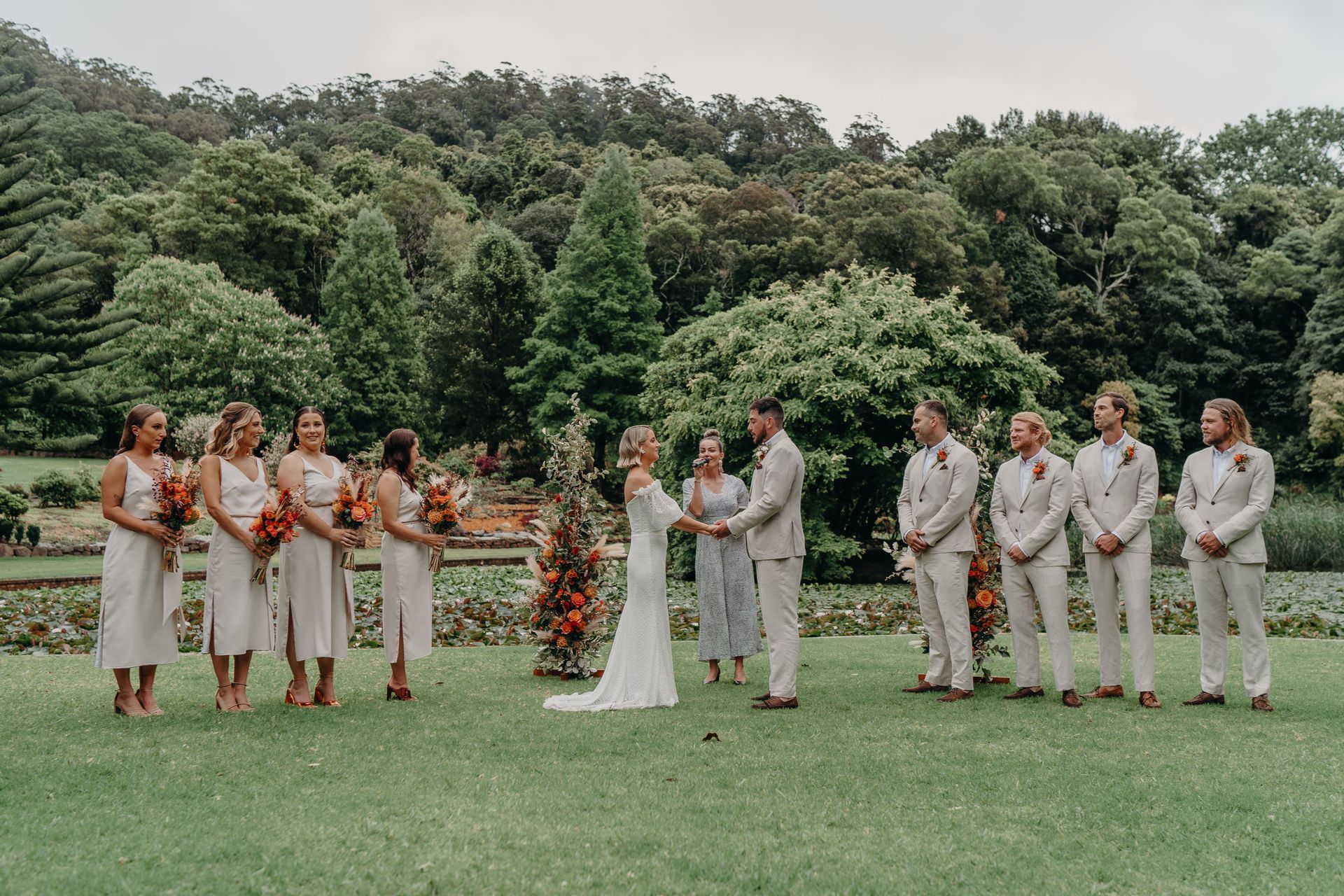 Wedding ceremony outdoors. Bride and groom holding hands, bridesmaids, groomsmen, lush greenery.