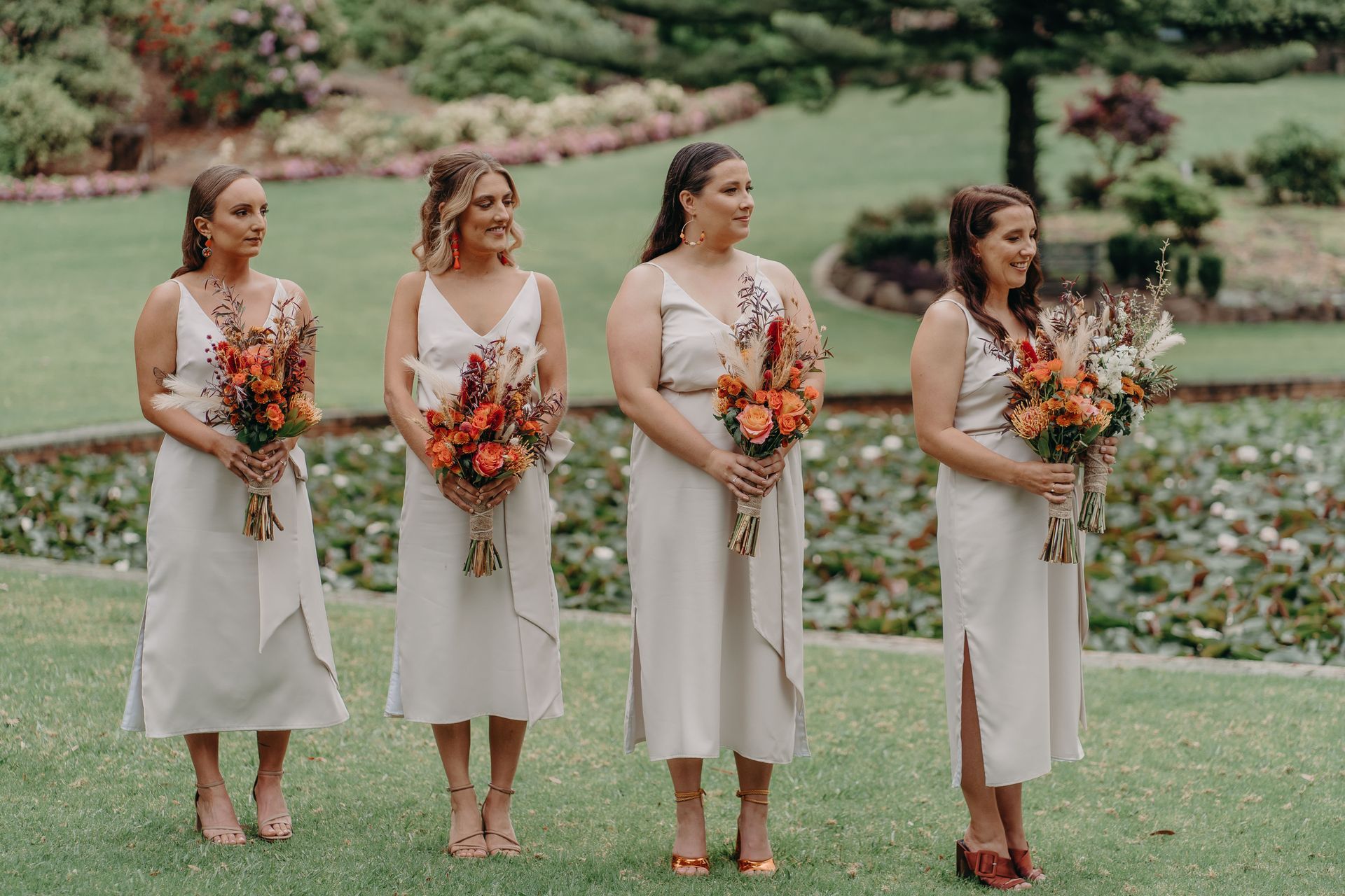 Four bridesmaids in cream dresses hold bouquets in a garden, smiling at a wedding.