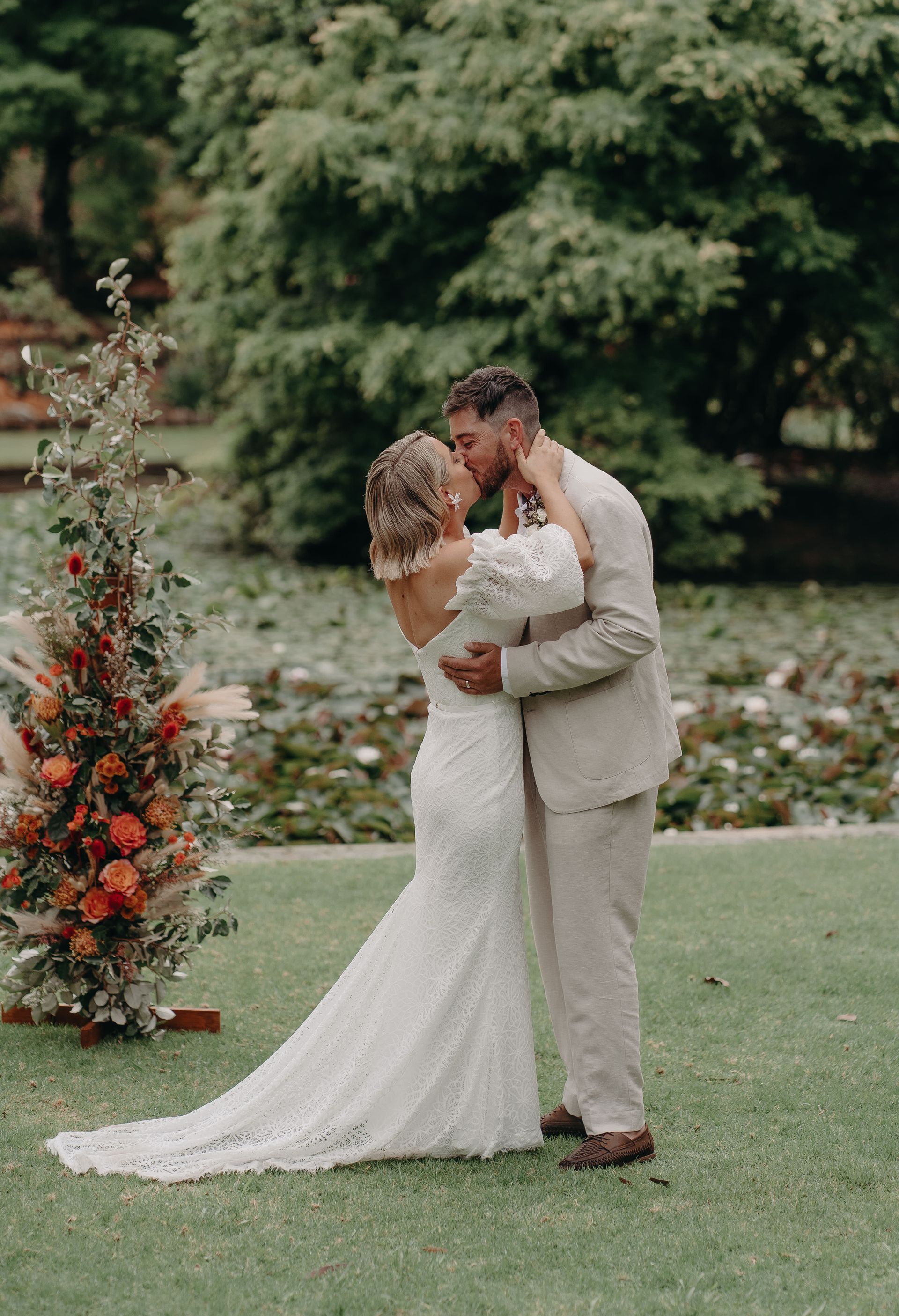 Newlyweds kissing in a garden. The bride wears a lace gown; the groom, a tan suit. Ceremony backdrop.