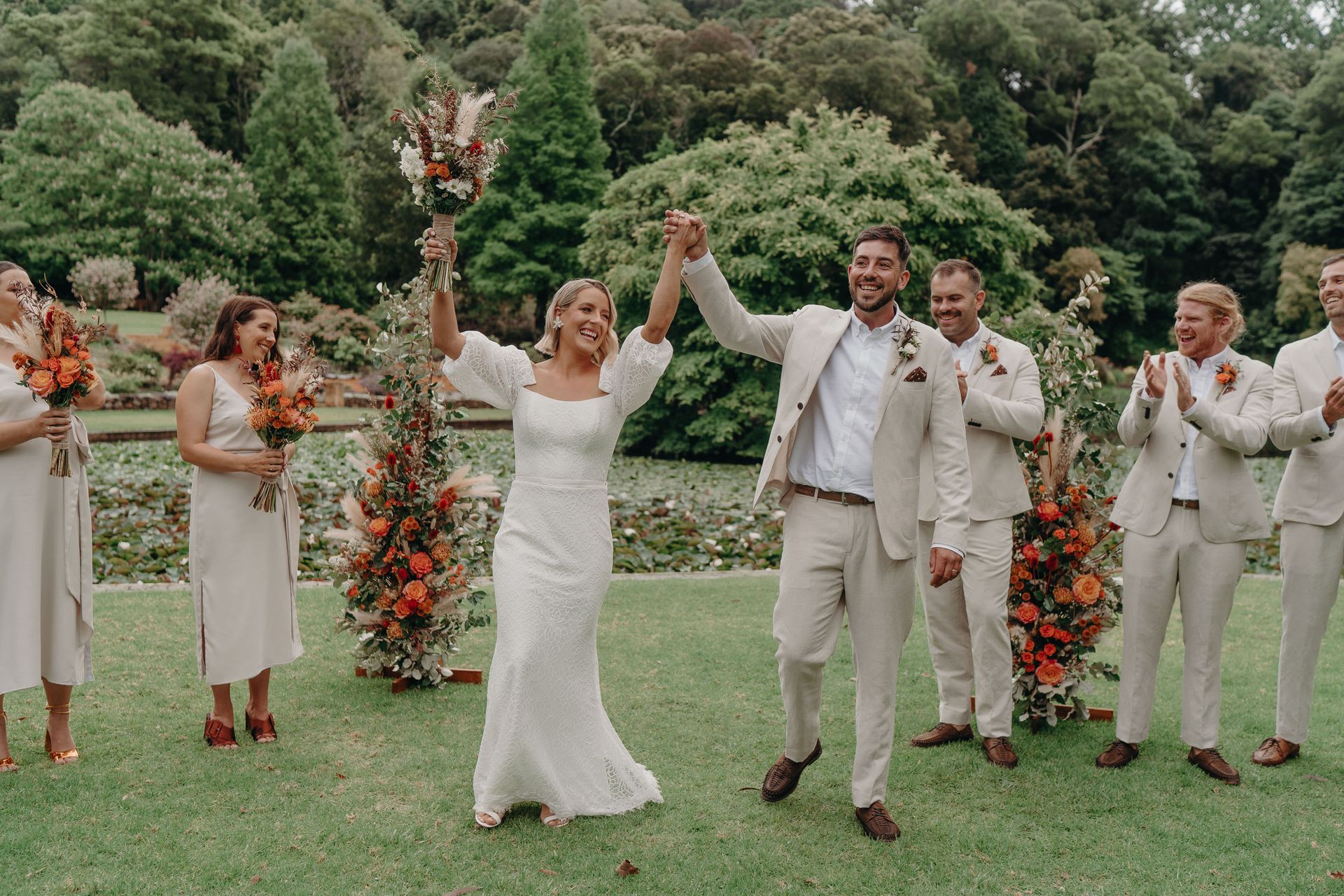 Newly married couple raising hands, walking on grass with wedding party applauding in garden.