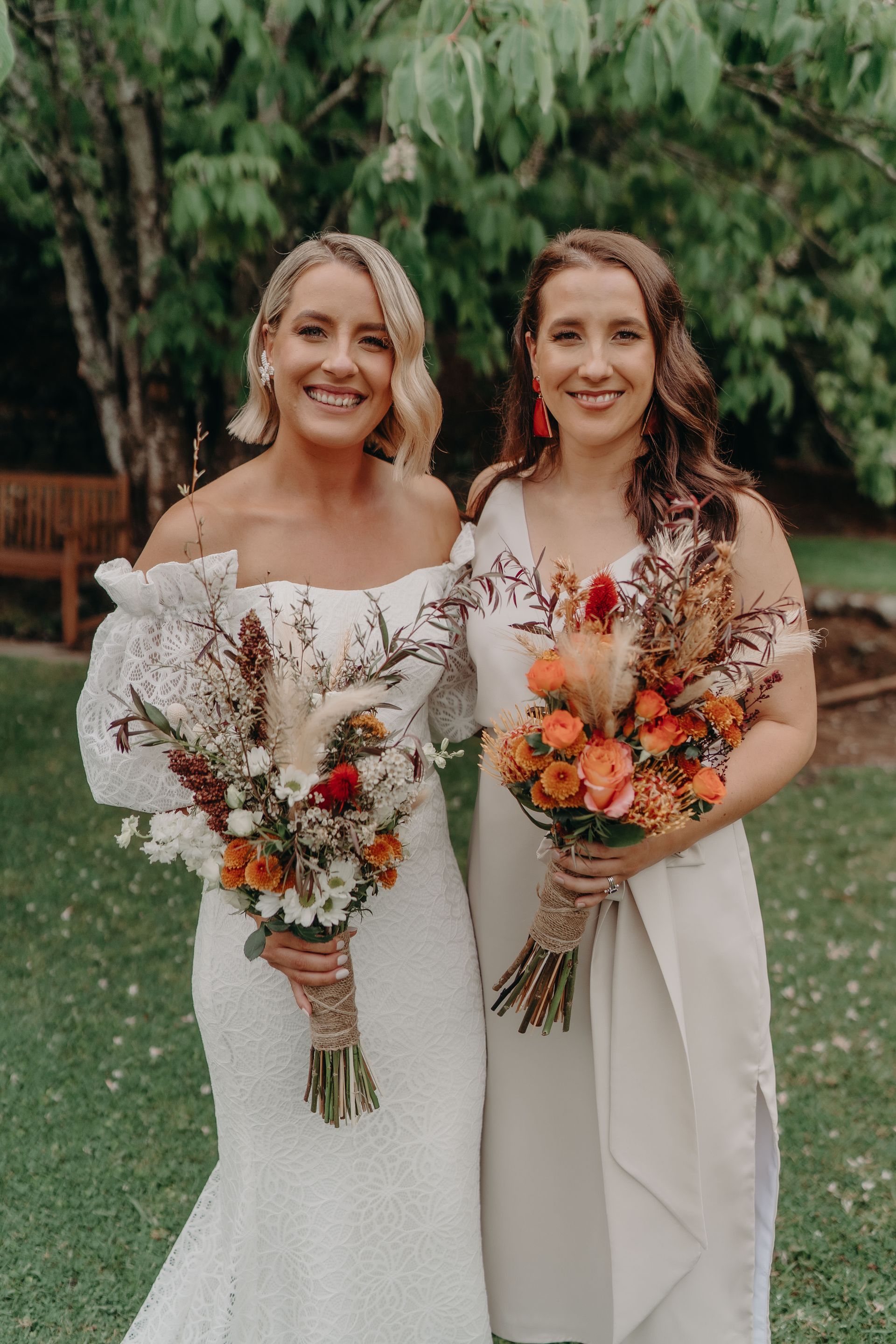 Two smiling women in white dresses, holding bouquets outdoors.