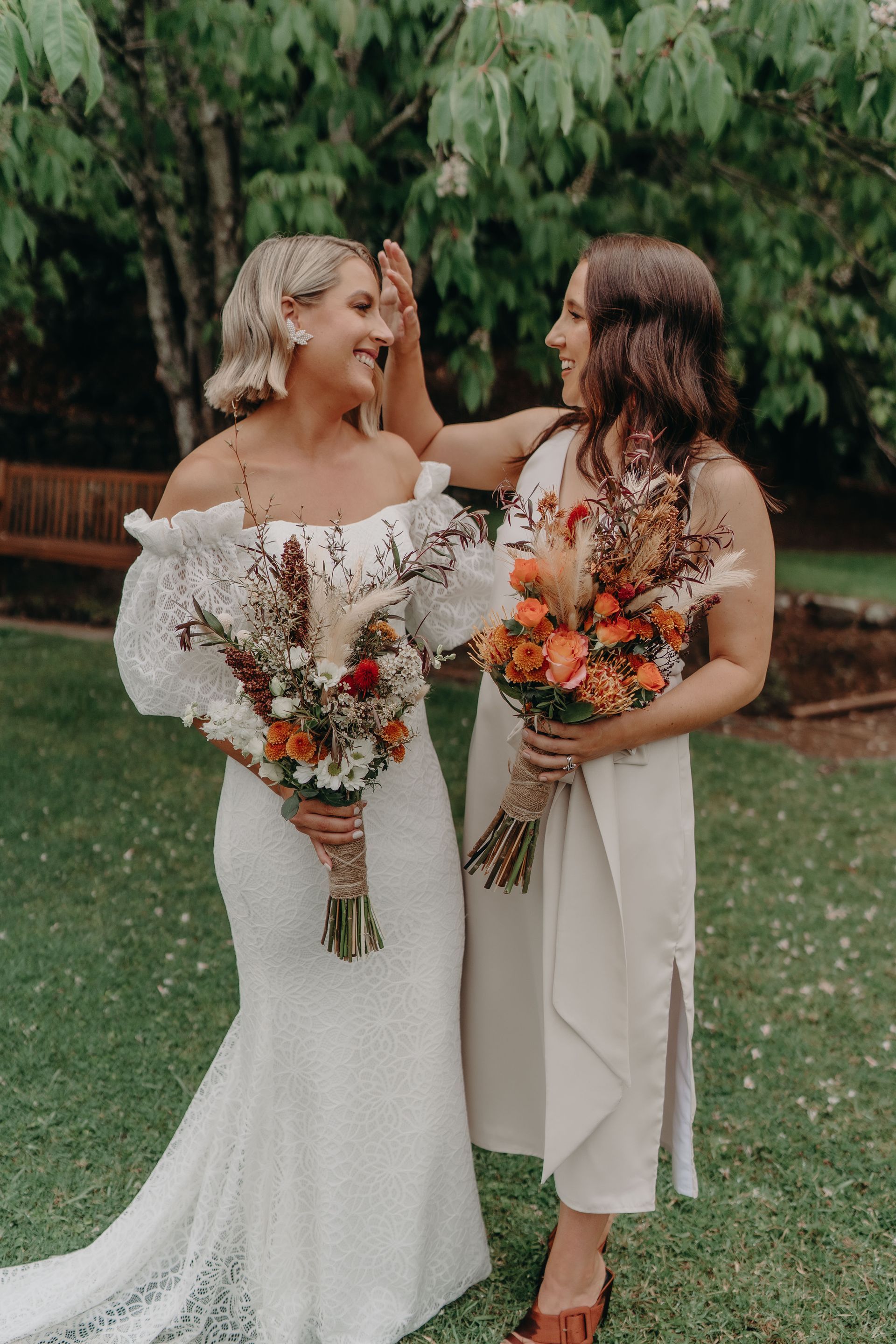 Bride in white dress with bridesmaid; holding bouquets, smiling outdoors.