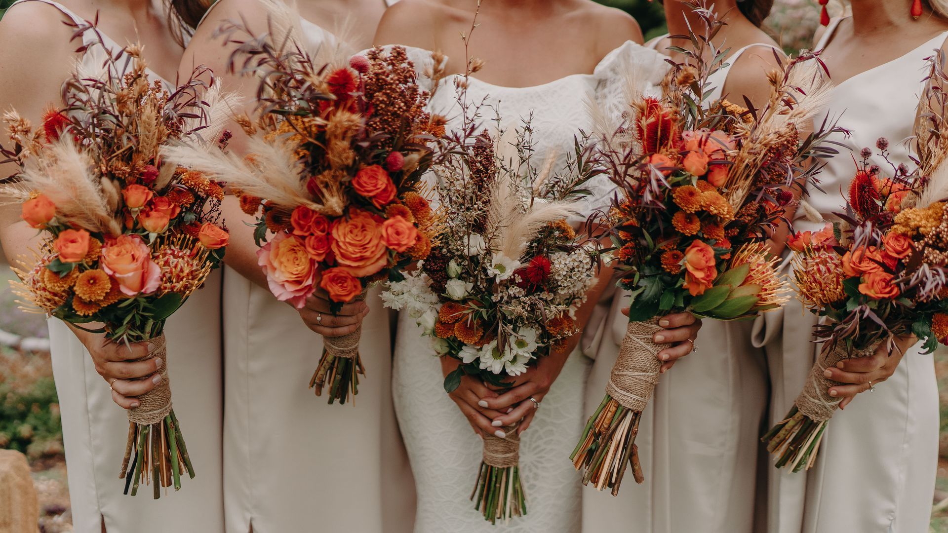 Bridesmaids in cream dresses holding autumn-toned bouquets with orange and brown flowers.