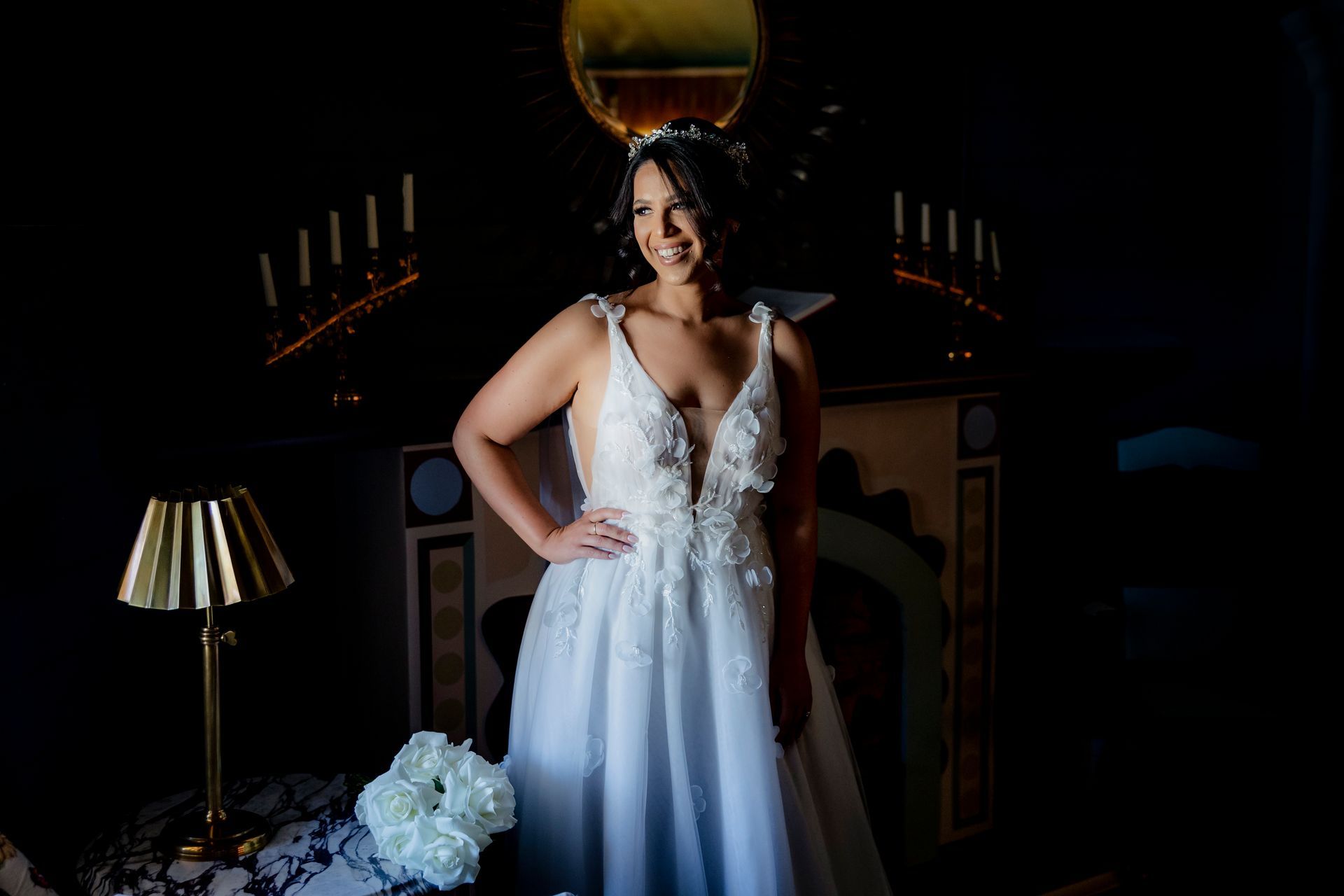 Bride in a white wedding dress stands near a fireplace, smiling with hand on hip.