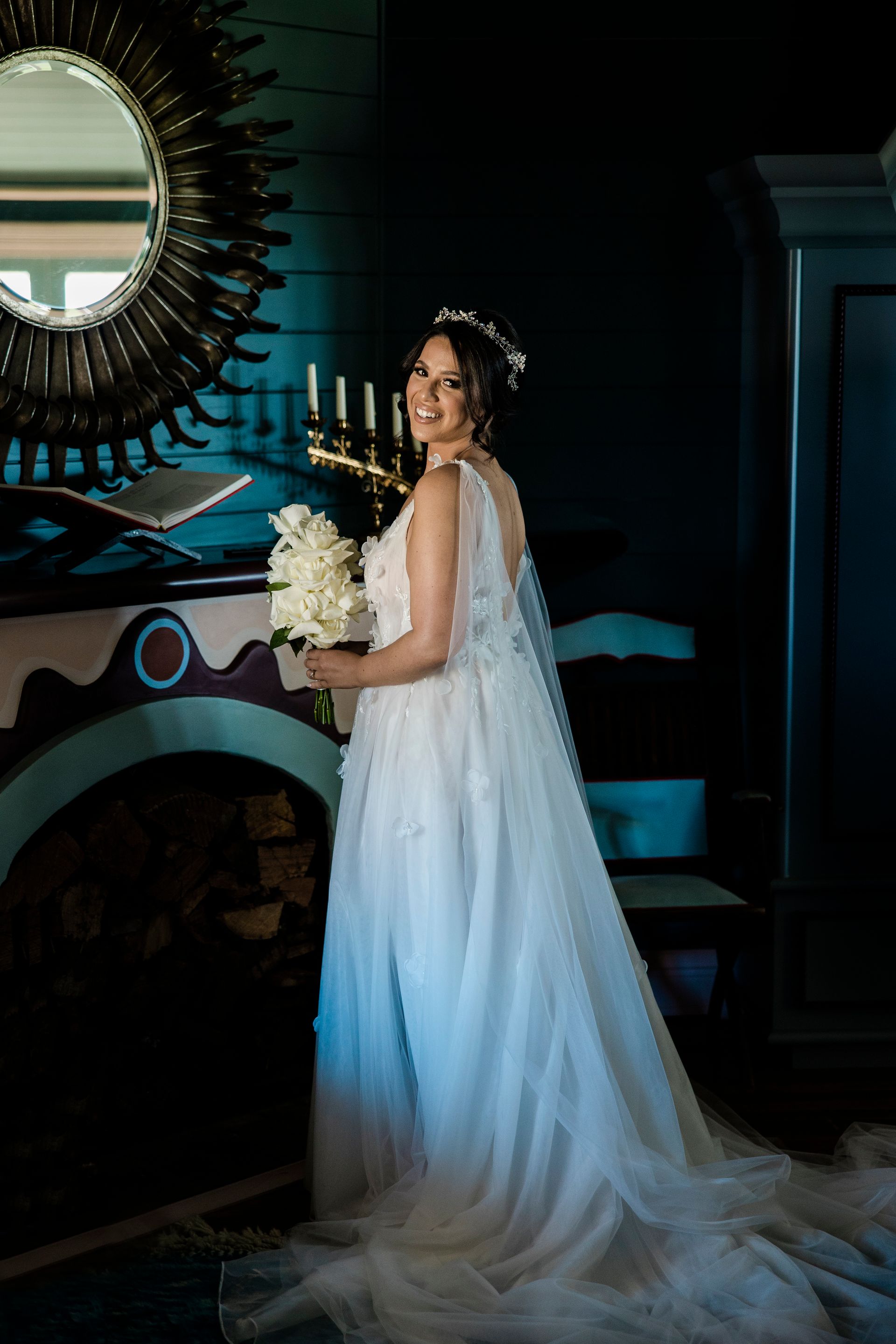 Bride in a white gown and veil, holding flowers, smiling in front of a mirror and fireplace.