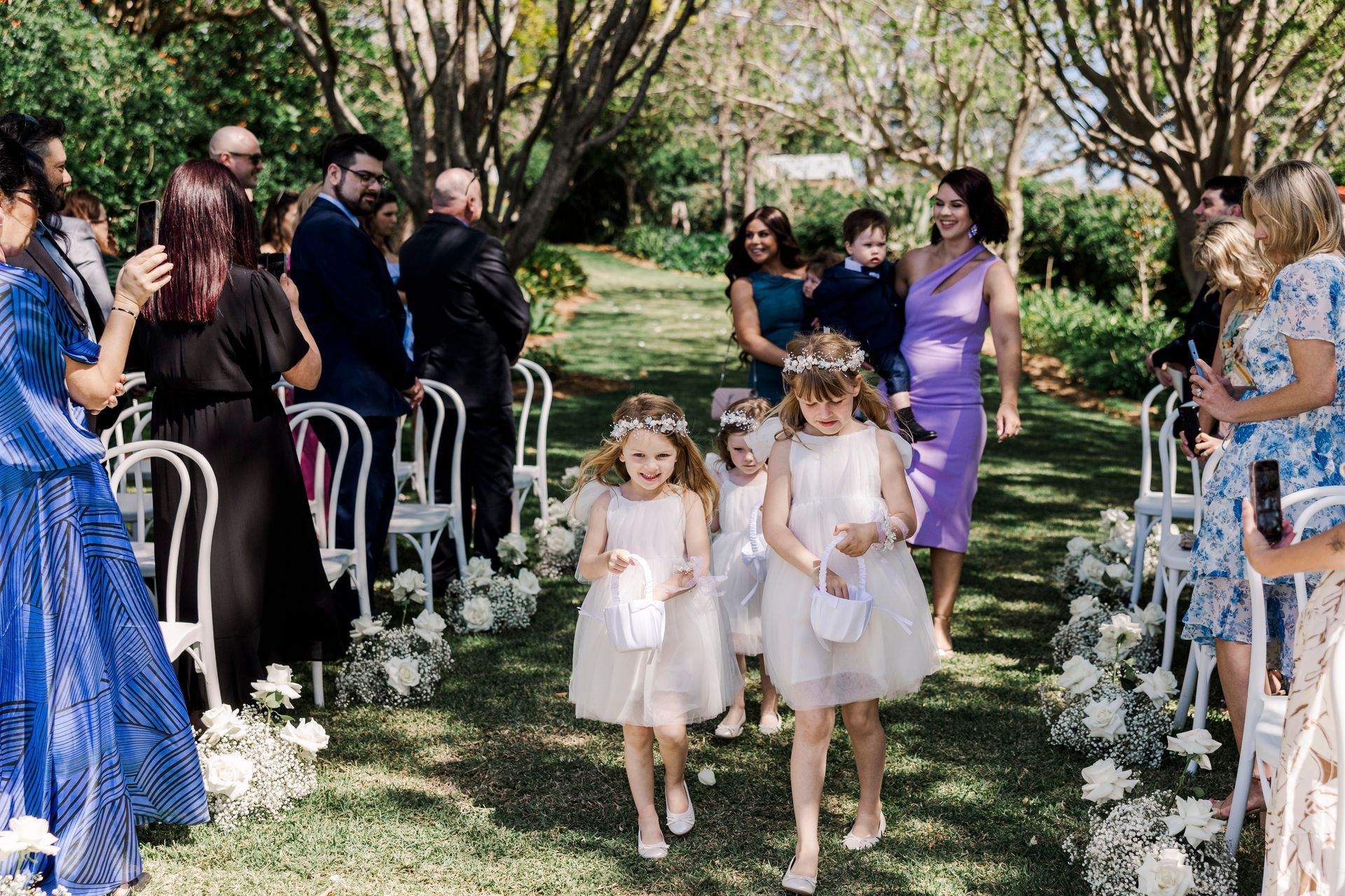 Flower girls in white dresses walk down a grassy aisle during an outdoor wedding ceremony.