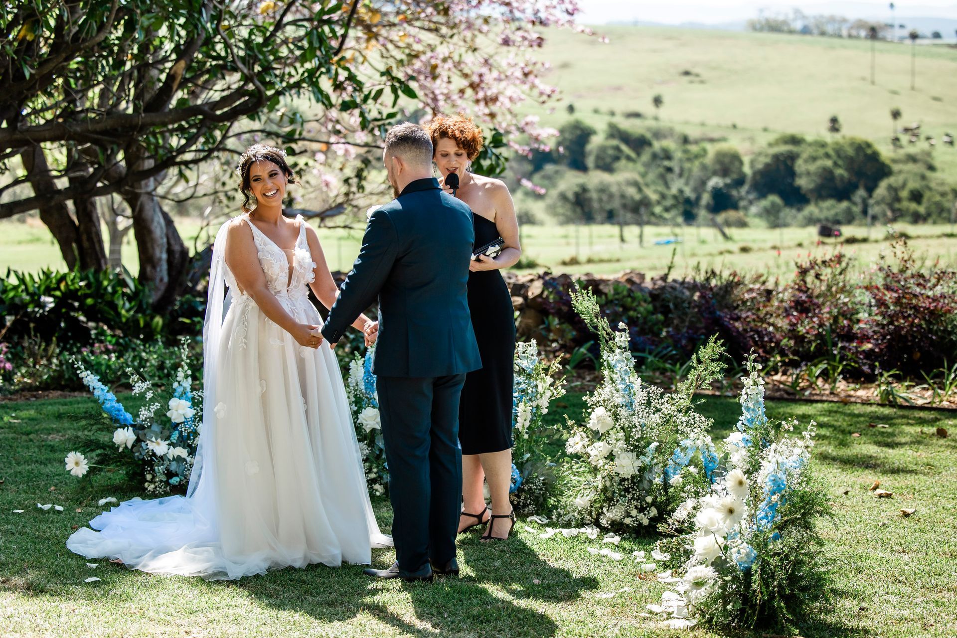 Couple holding hands at outdoor wedding ceremony; floral arch, green landscape.