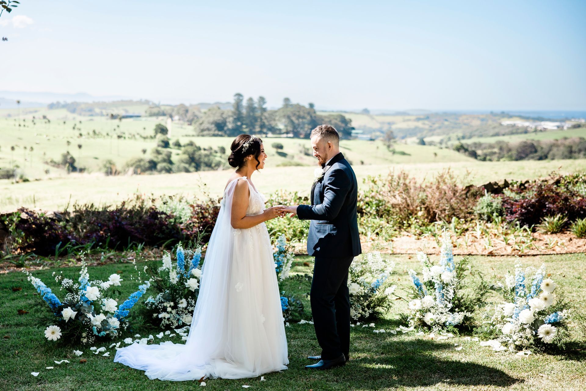 Bride and groom exchanging rings at outdoor wedding ceremony with floral decorations, scenic landscape in background.