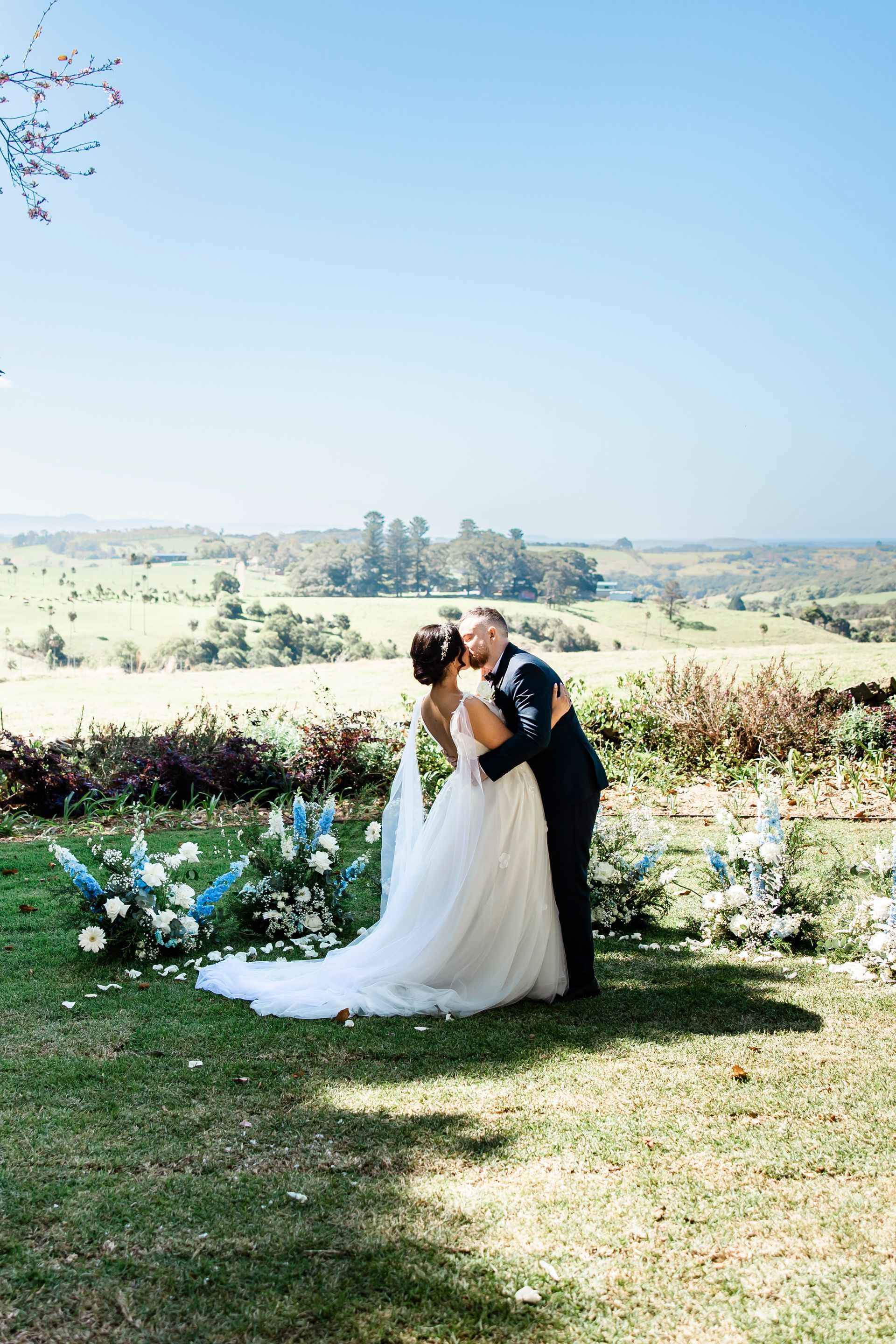 Couple kissing during outdoor wedding ceremony, with floral arch, fields, and blue sky.
