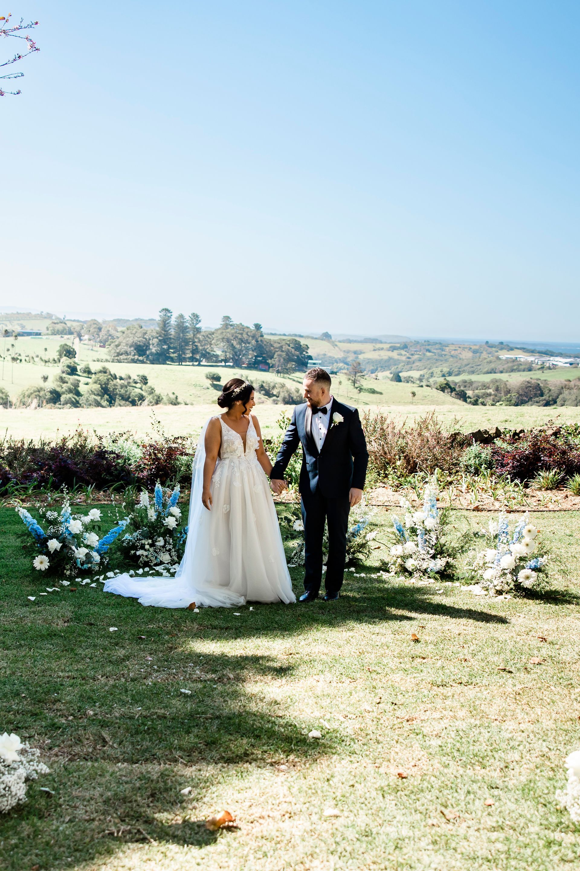 Bride and groom holding hands, standing outdoors in front of floral arrangements and a scenic landscape.