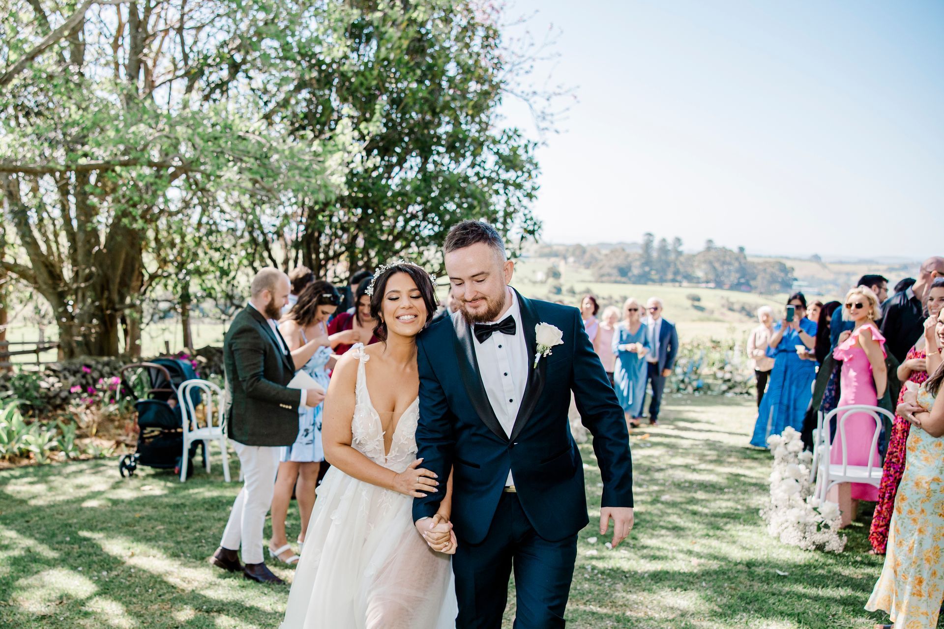 Newlyweds walk hand-in-hand down an outdoor aisle, smiling. Guests stand nearby, celebrating.