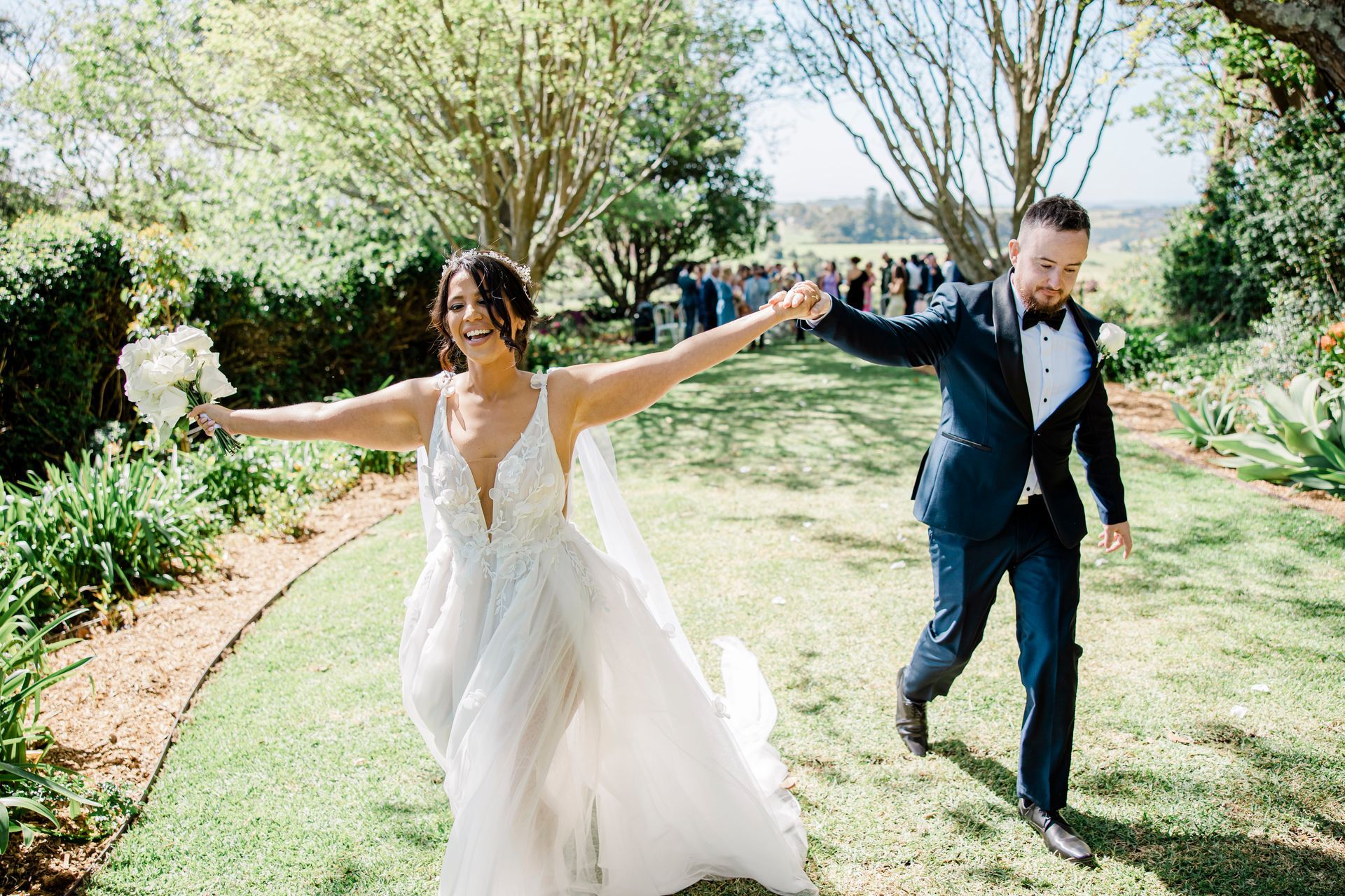 Newlyweds run down a grassy path. The bride wears a white gown, holding flowers. The groom wears a blue suit.