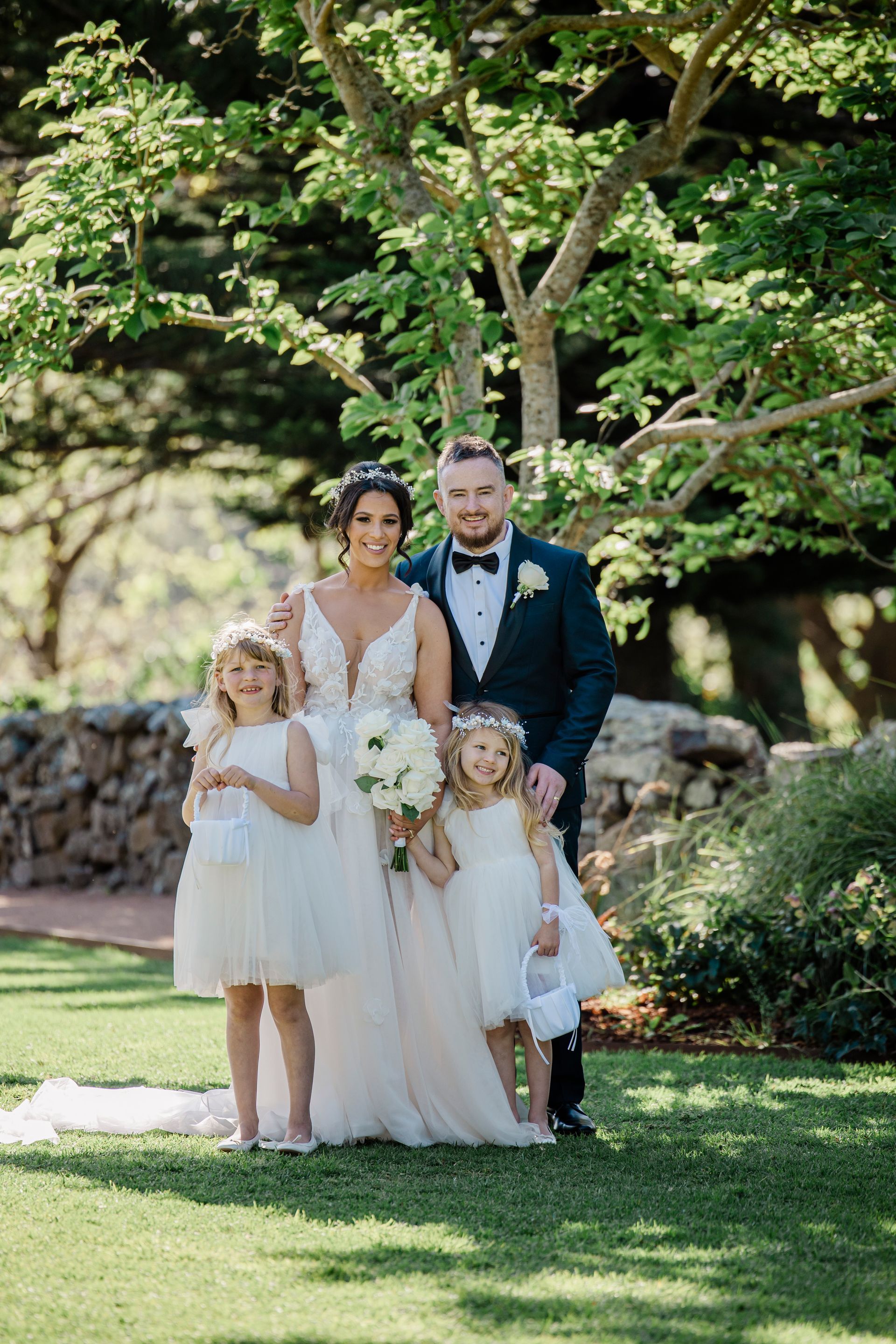 Wedding couple with flower girls pose in a garden. The bride wears white, groom in a dark suit.