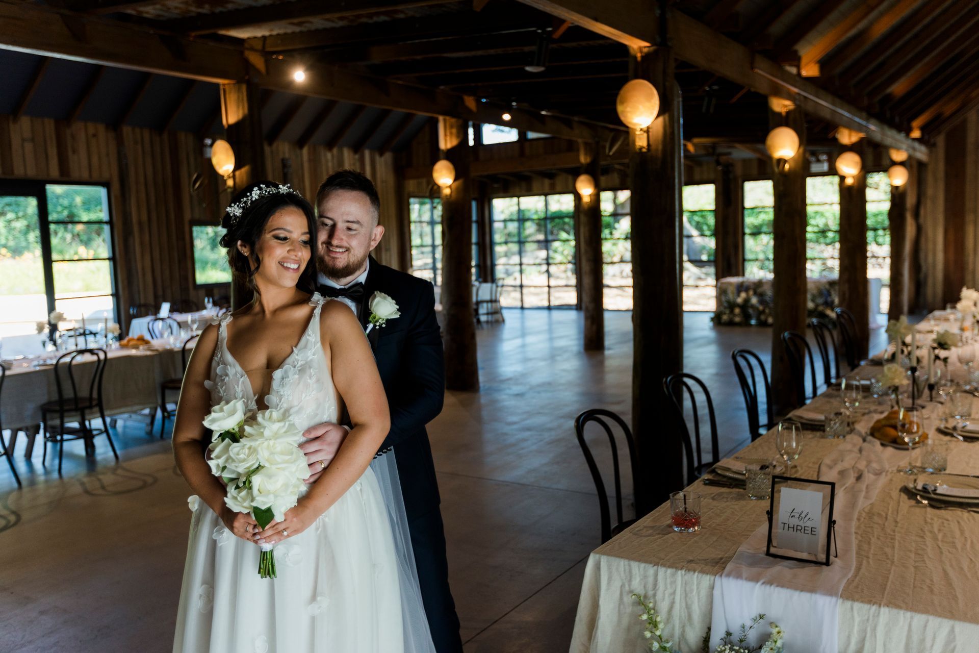 Wedding couple inside a barn. Woman in white dress holds bouquet; man in tuxedo. Tables set, string lights.