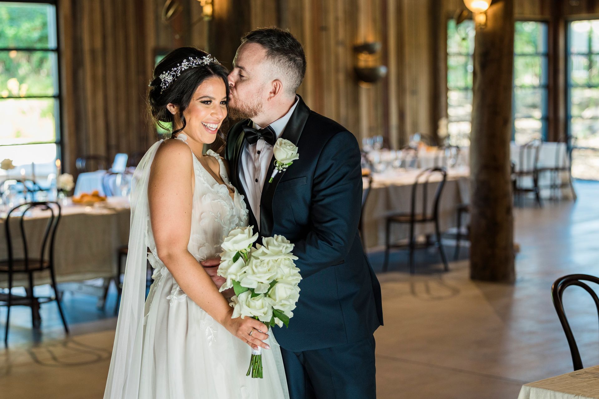 Wedding couple: man kisses woman's forehead, holding flowers. Inside, wood-paneled room with tables and chairs.