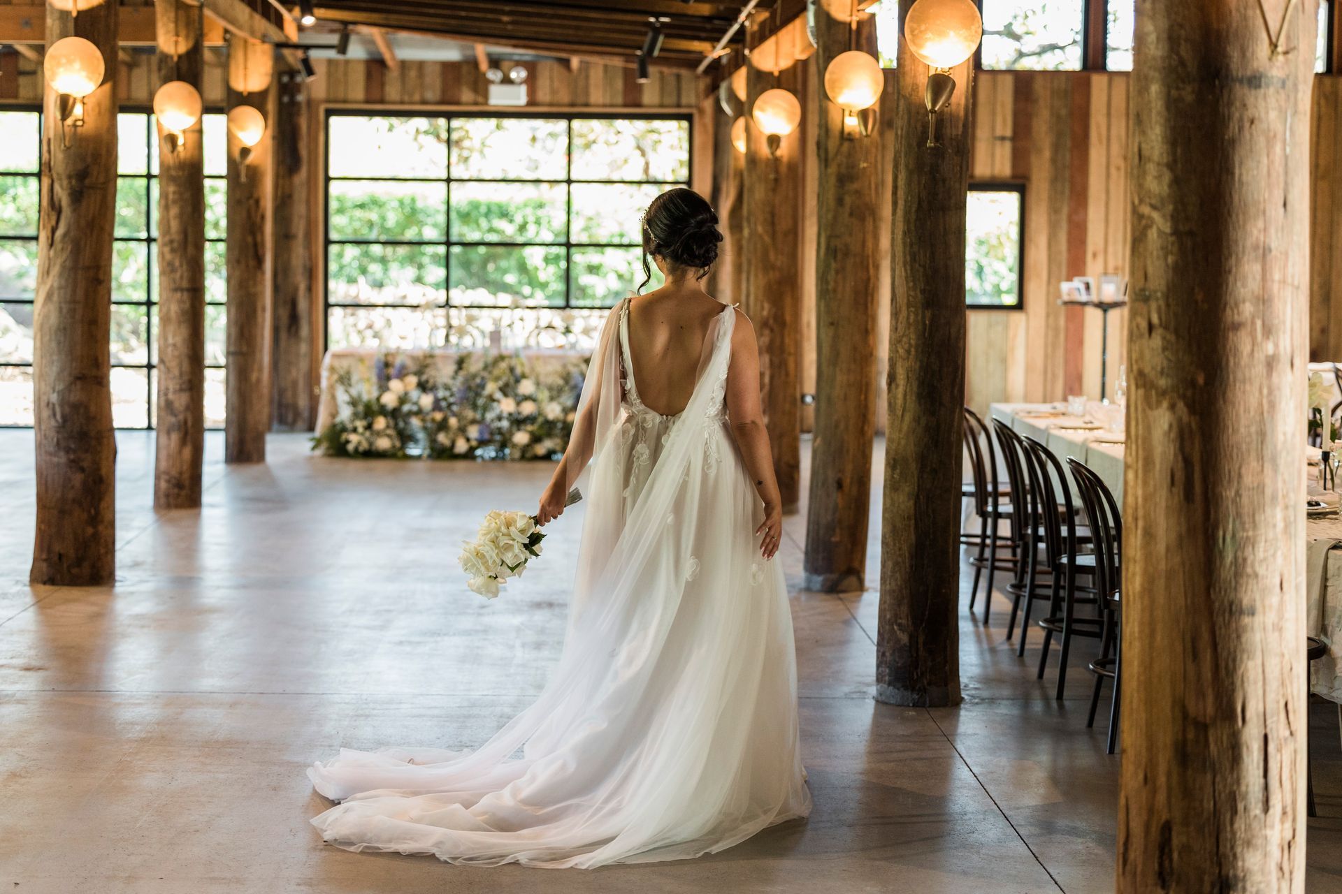 Bride in flowing white dress holding bouquet, walking in rustic venue, with wood beams and large window.