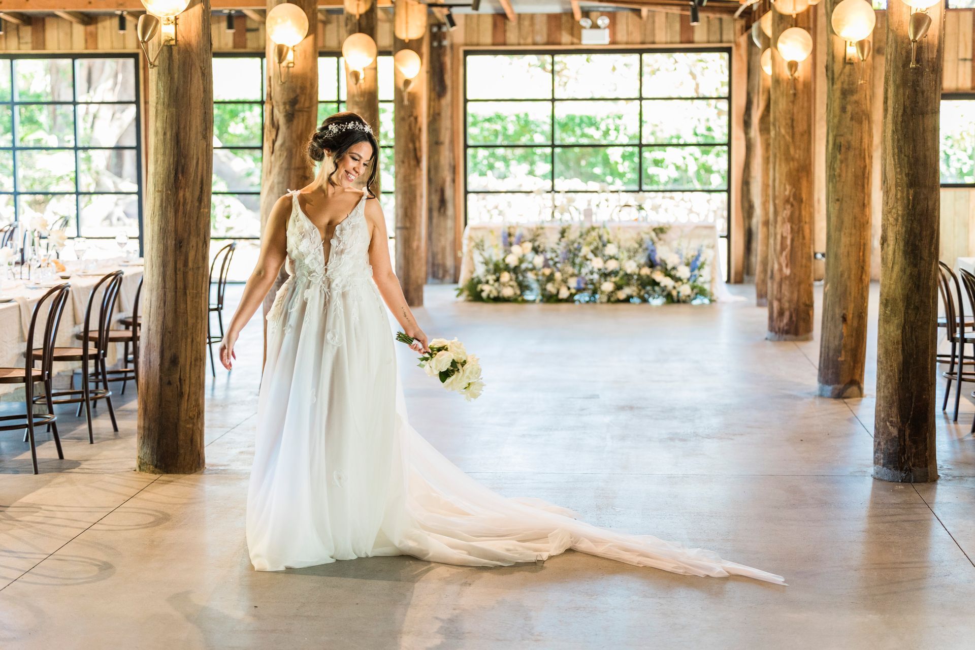 Bride in a wedding dress smiles, holding bouquet. Inside a wooden venue with a long train.