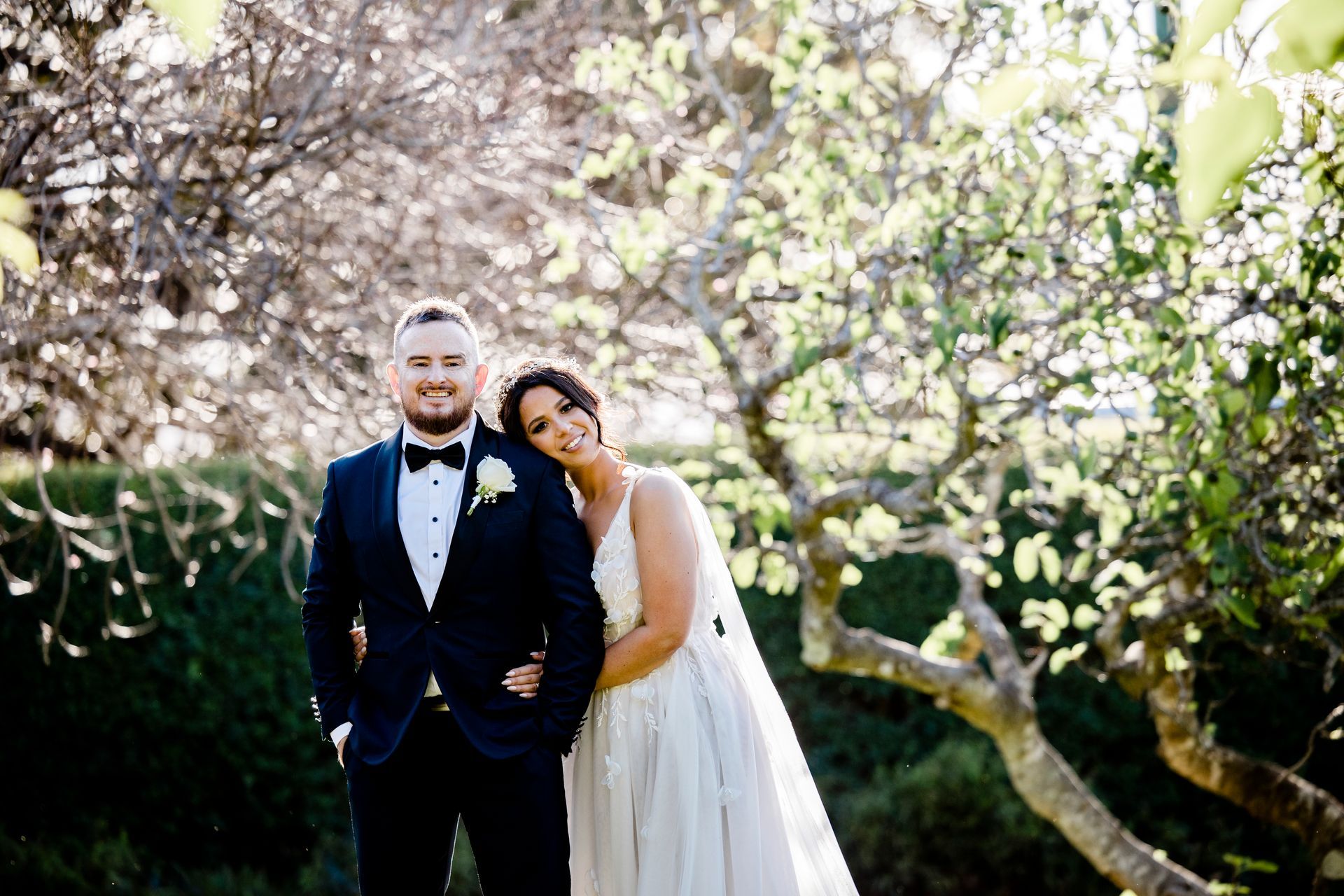 Bride and groom pose outdoors in garden; man in dark suit, woman in white dress, both smiling.