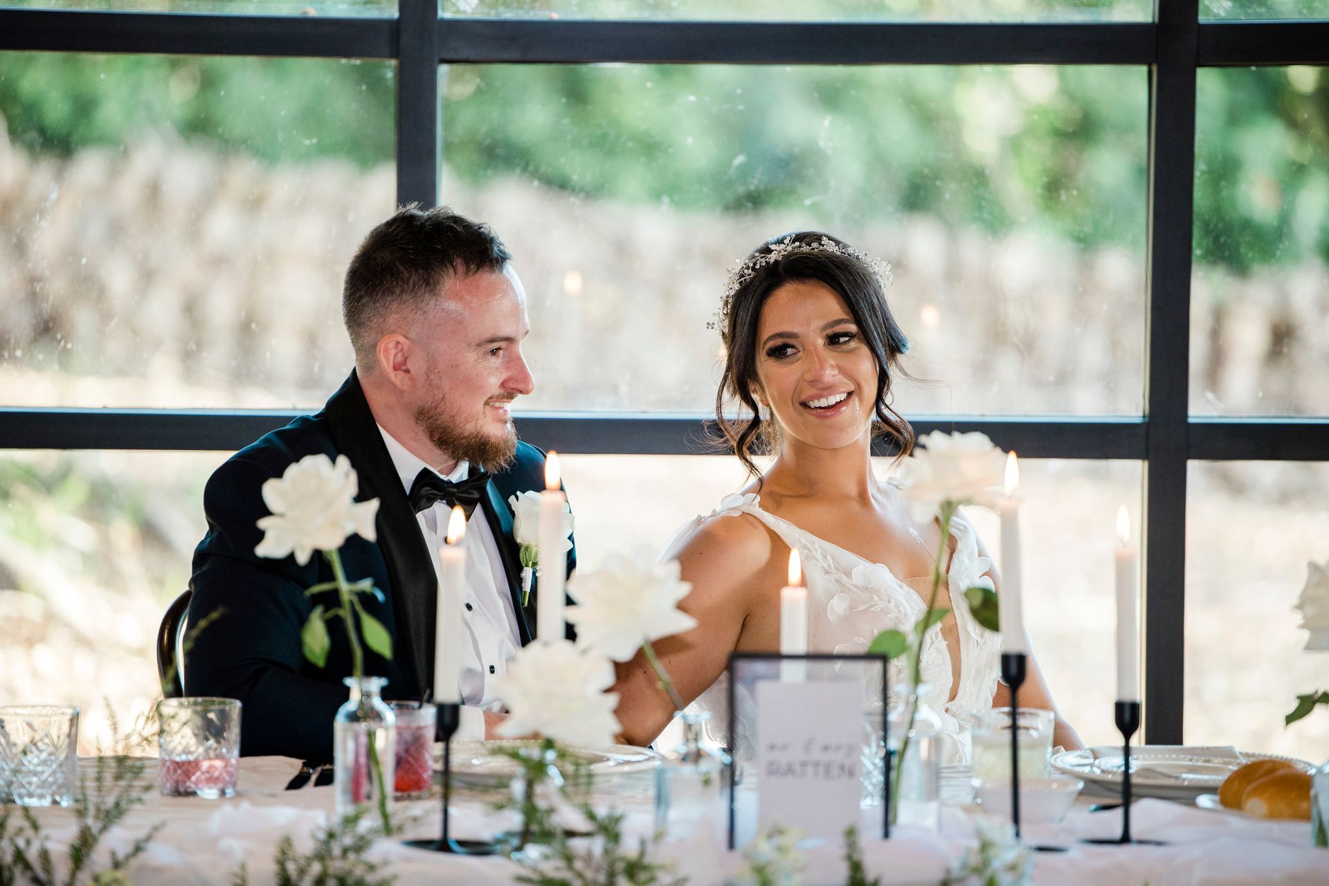 Bride and groom at a wedding reception table; smiling and looking toward the same direction.