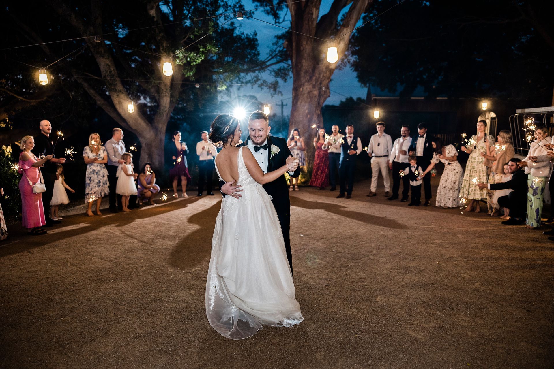 Couple dances at wedding, surrounded by guests holding sparklers, beneath string lights. Outdoor setting, night.