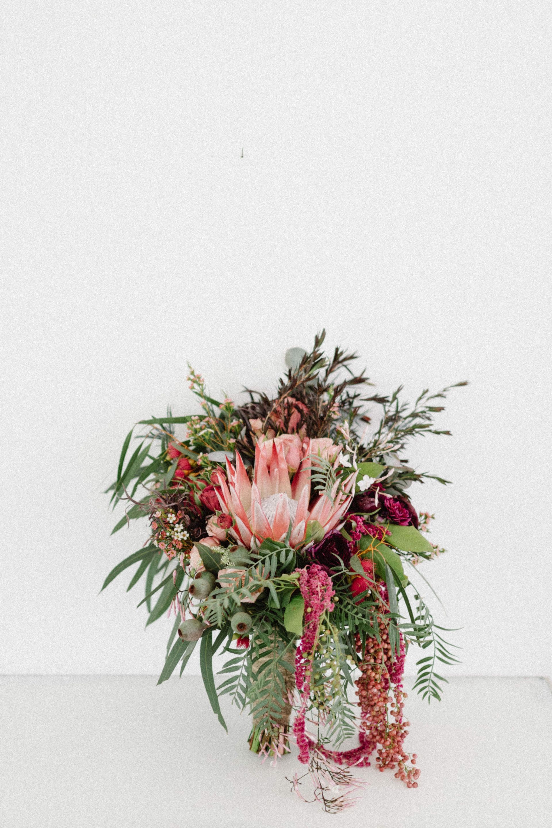 Protea flower bouquet against a white wall, featuring pink, red, and green foliage.