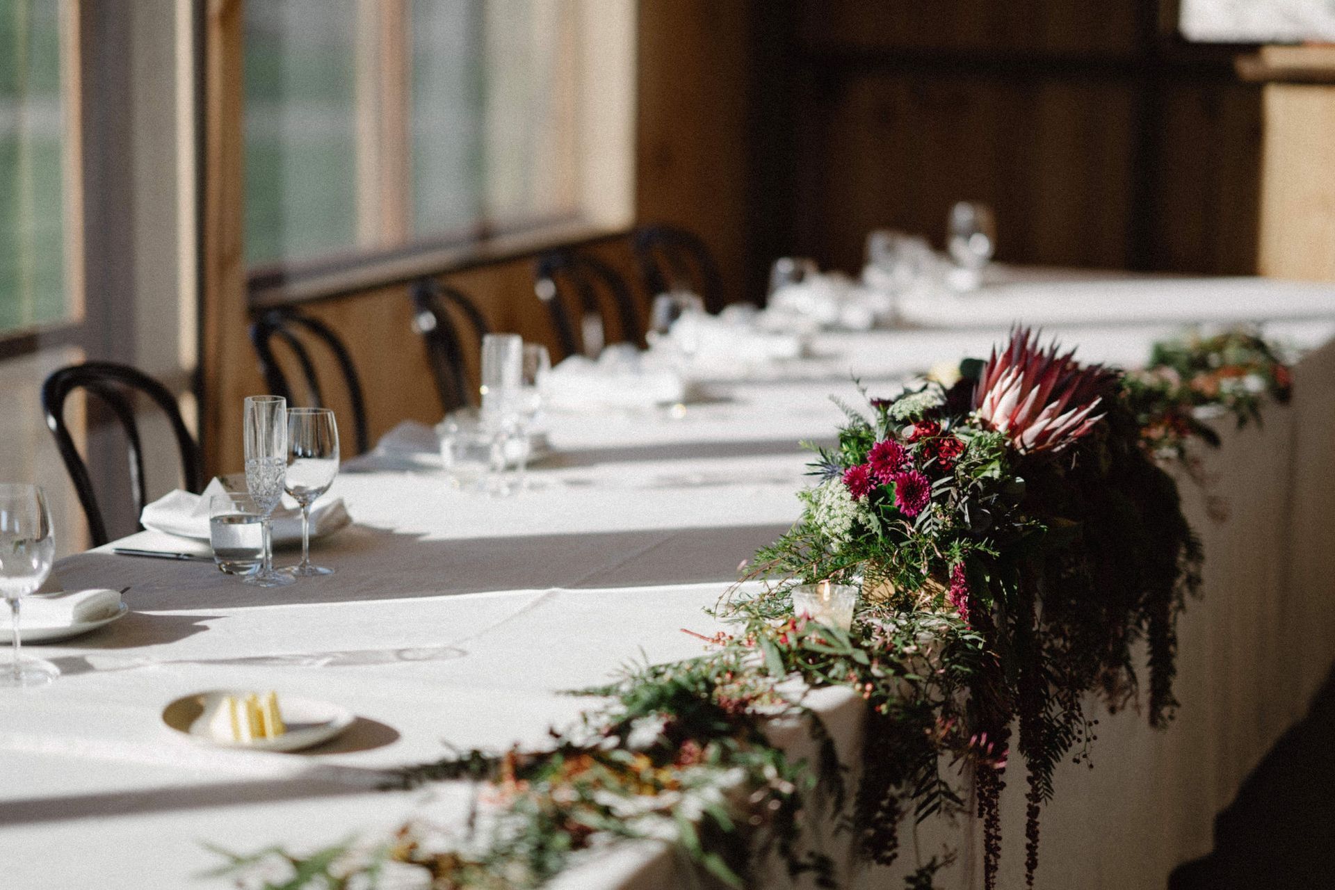 Wedding reception table set with flowers, glasses, and plates in a sunlit room.