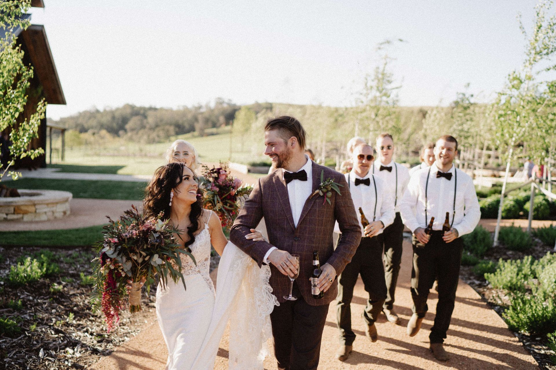 Bride and groom walking, laughing with wedding party. Sunny outdoor setting.